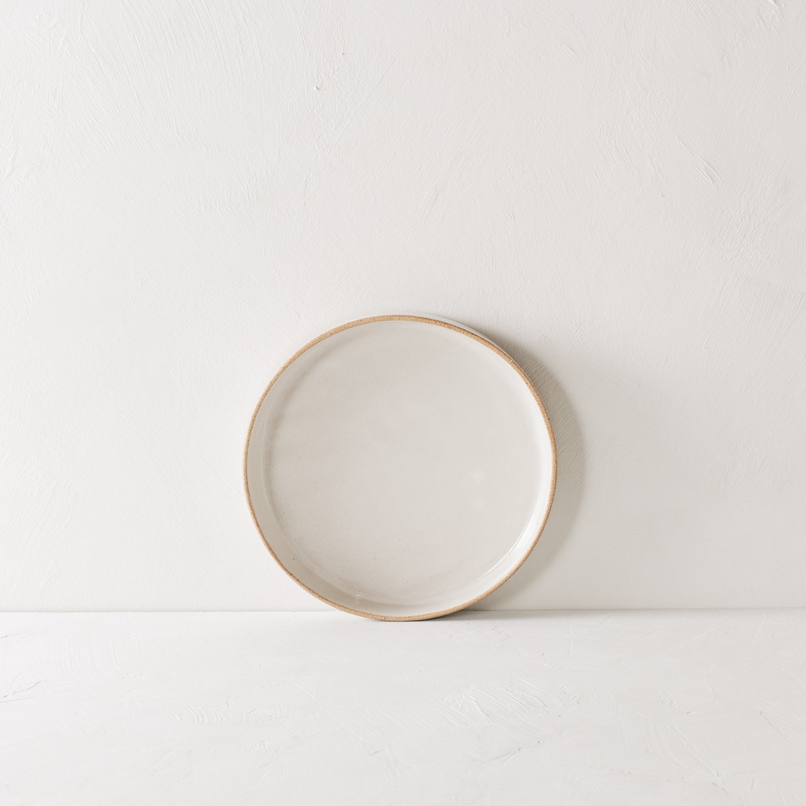 Minimal white dinner dish leaning against the backdrop. Exposed stoneware rim can be seen. Handmade ceramic dinner dish, designed and sold by Convivial Production, Kansas City ceramics.