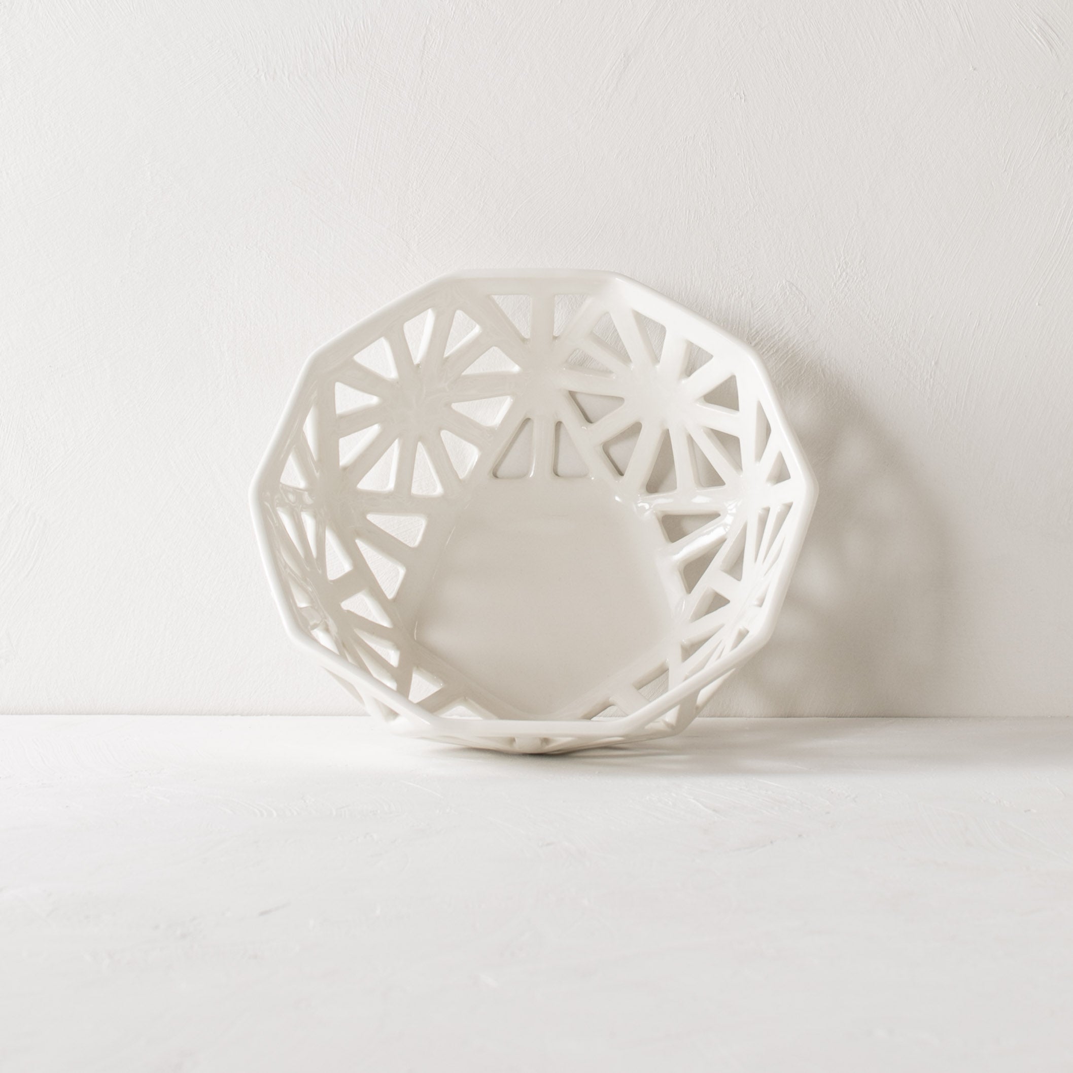 White geometric fruit bowl. Pentagon carved design with additional carved designs in the shape of slim right angle triangles. Staged on a white plaster table top and a white plaster textured wall. Fruit bowl is leaning against the wall upright to show the full overhead view. Designed and sold by Convivial Production, Kansas City Ceramics