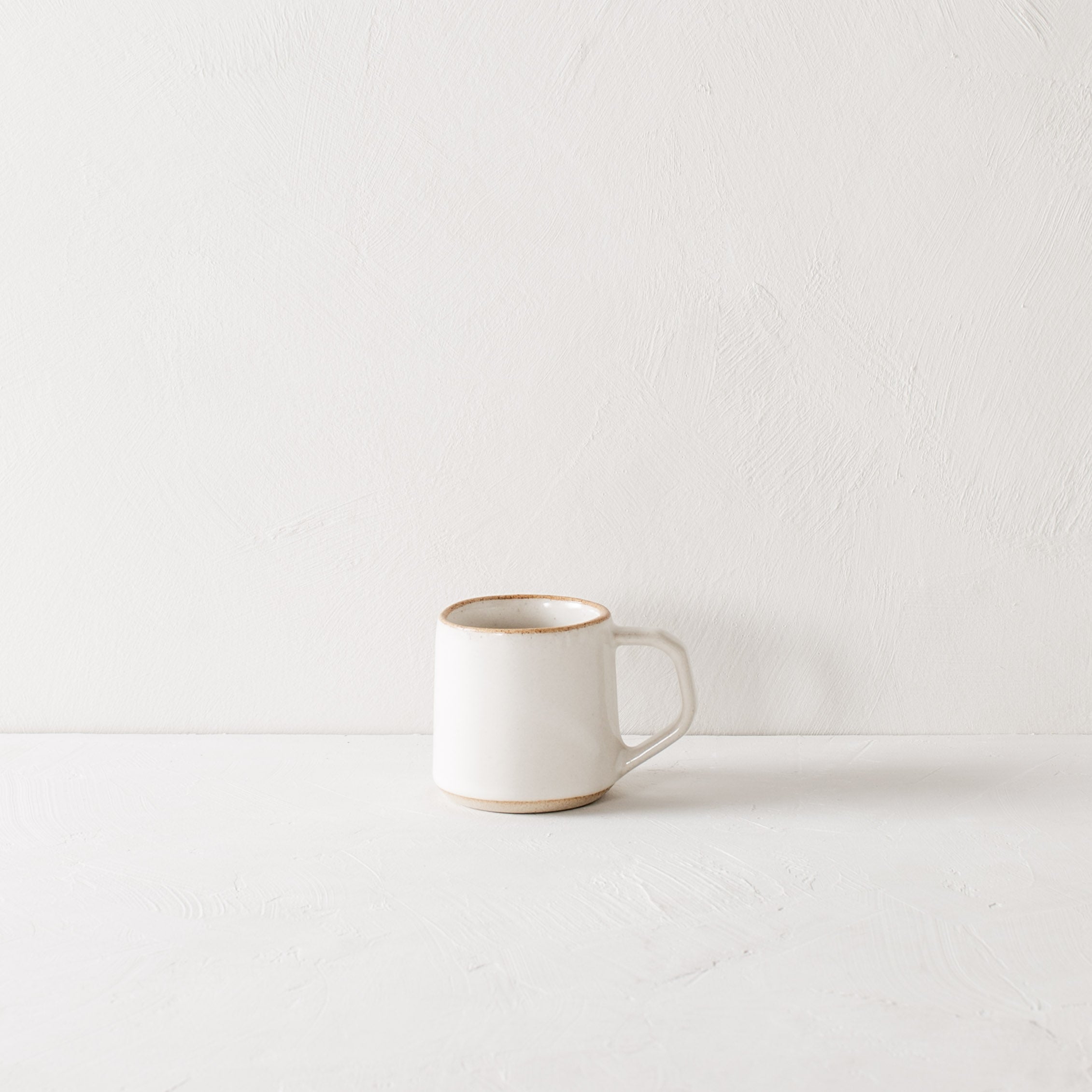 White minimal ceramic mug is staged on a white textured table top and white textured back drop. Mug has an exposed warm stoneware on the rim as well as the base. Handle is geometric in shape but still rounded. Convivial Production, Kansas City Ceramics. Handmade ceramic mug.  