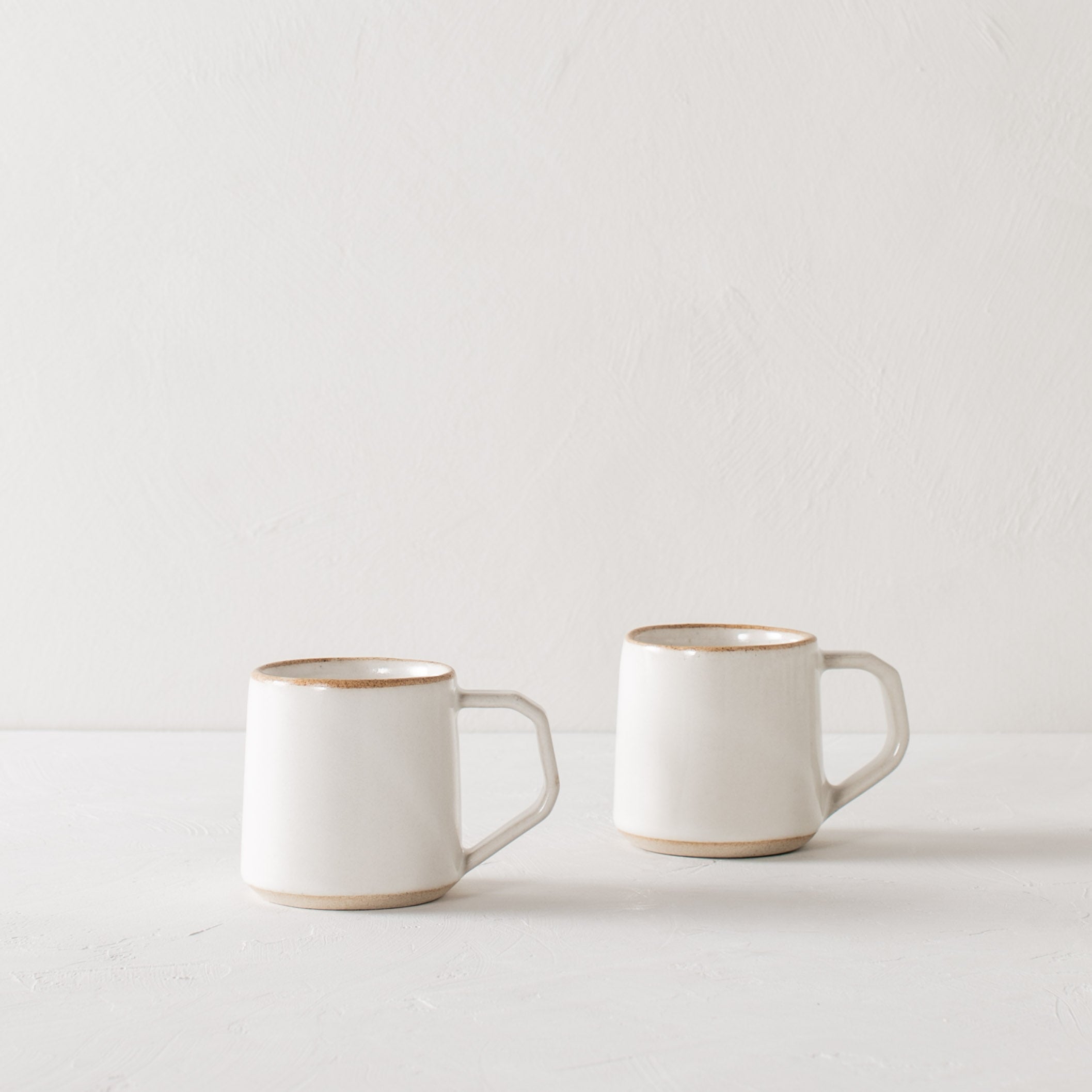 Pair of white minimal ceramic mug is staged on a white textured table top and white textured back drop. Mugs have exposed warm stoneware on the rims as well as the base. Handles are geometric in shape but still rounded. Convivial Production, Kansas City Ceramics. Handmade ceramic mug.  
