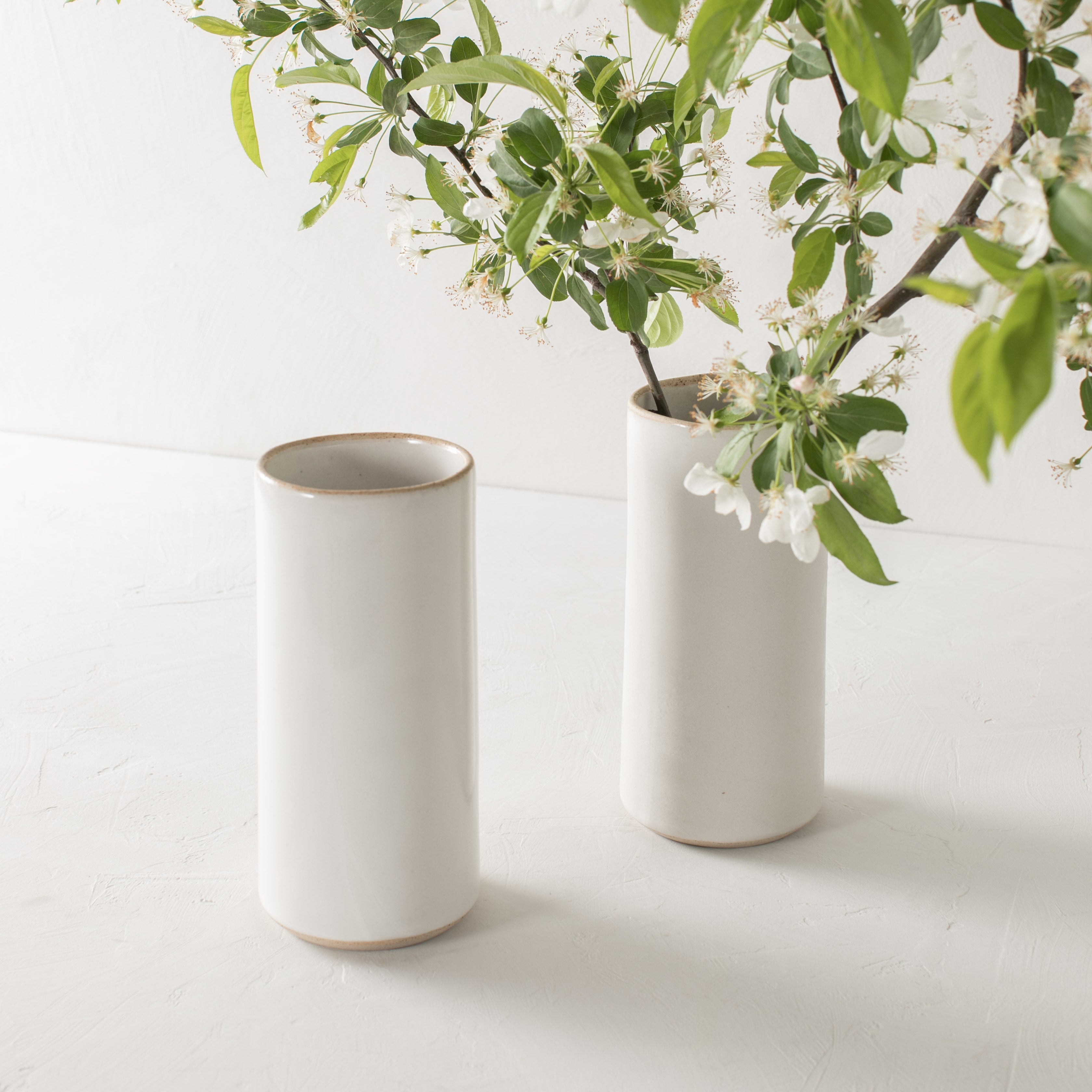 Two tall white glazed ceramic cylinder vases with an exposed stoneware rim and base. Detail up-close images focusing on vase holding a white blossom tree branch. Staged on a white textured plaster table and a white plaster textured wall. Designed and sold by Convivial Production, Kansas City Ceramics.