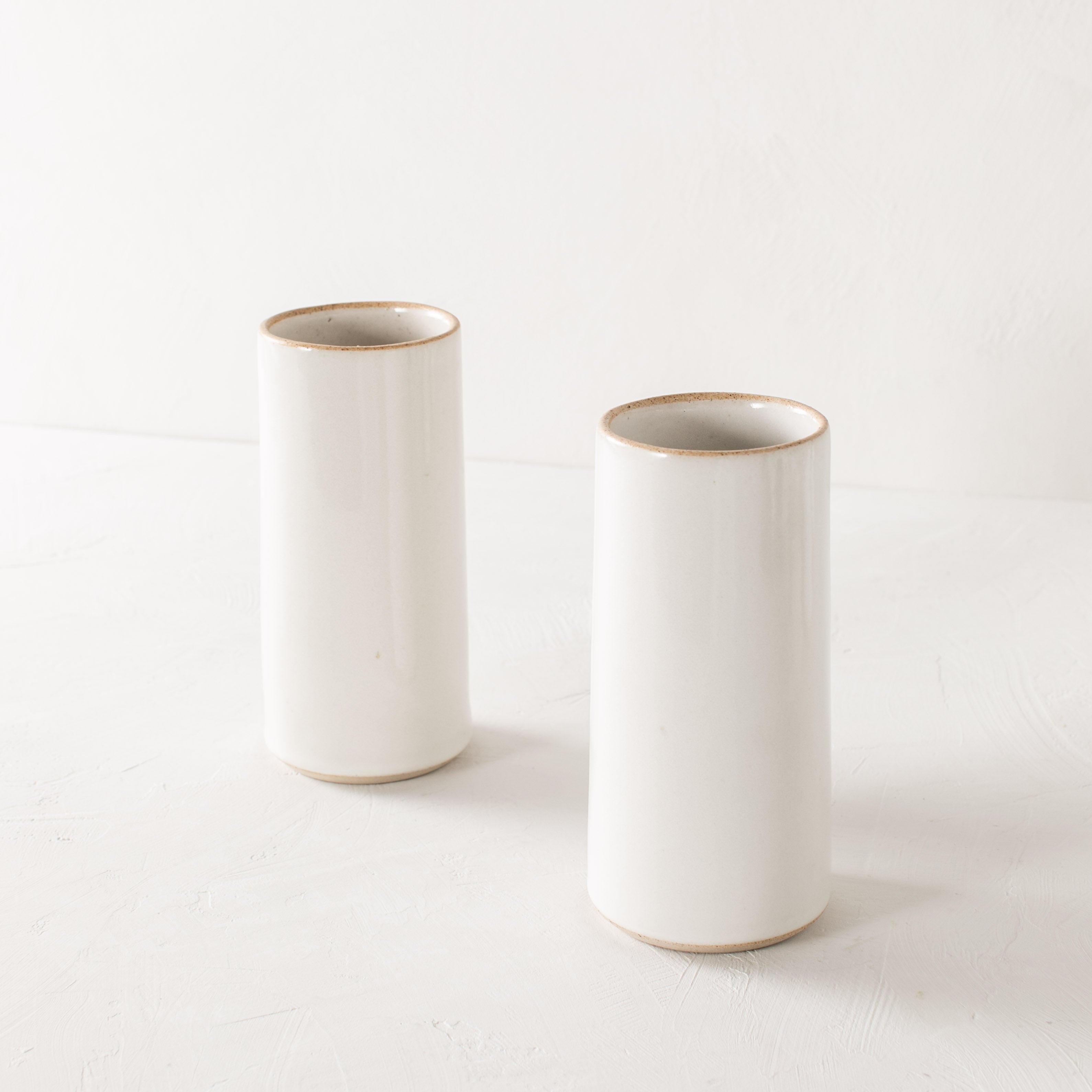 Two tall white glazed ceramic cylinder vases with an exposed stoneware rim and base. Staged on a white textured plaster table and a white plaster textured wall. Designed and sold by Convivial Production, Kansas City Ceramics.
