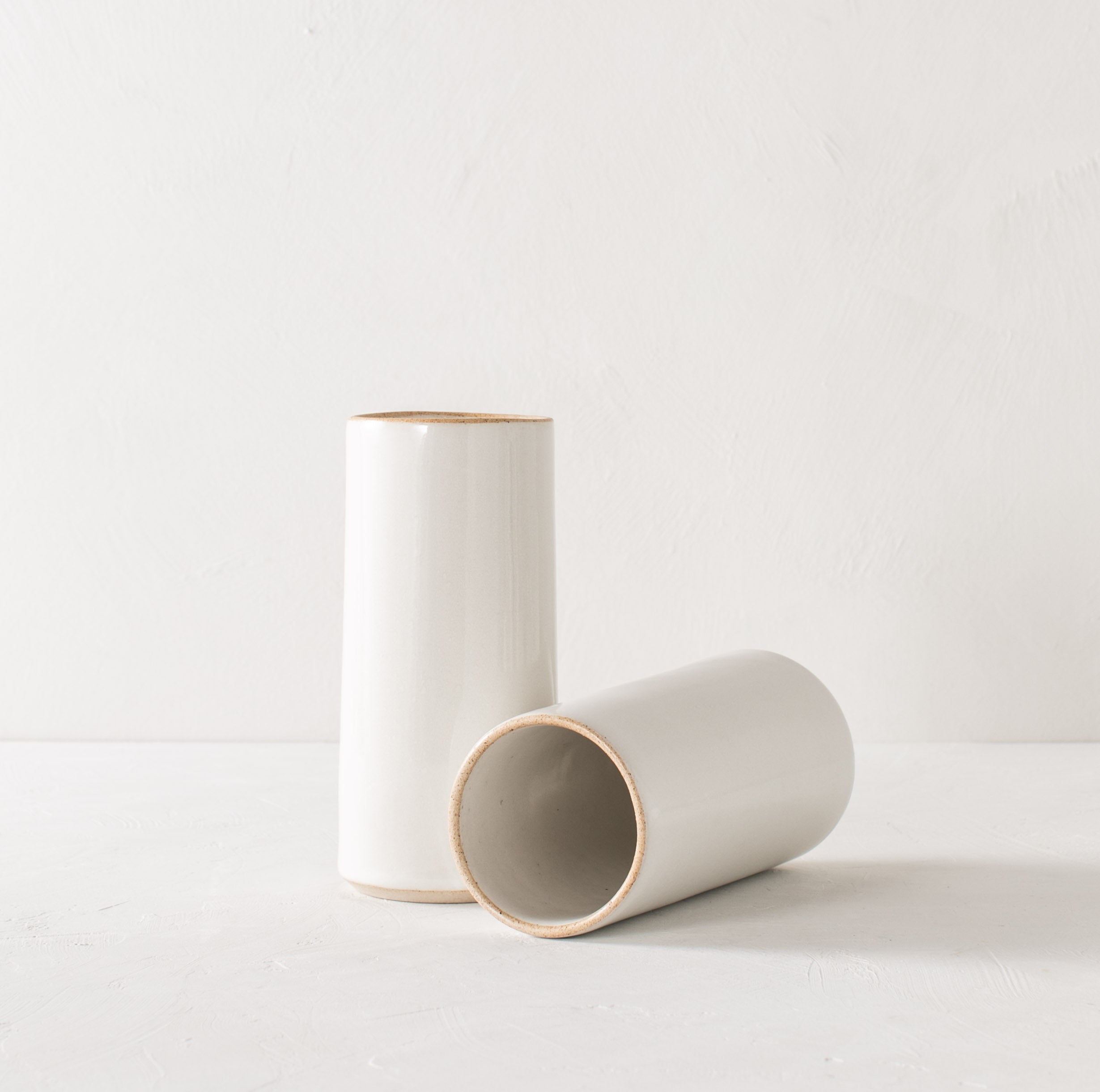 Two tall white glazed ceramic cylinder vases with an exposed stoneware rim and base. One standing up the other on its side leaning against the standing vase. Staged on a white textured plaster table and a white plaster textured wall. Designed and sold by Convivial Production, Kansas City Ceramics.
