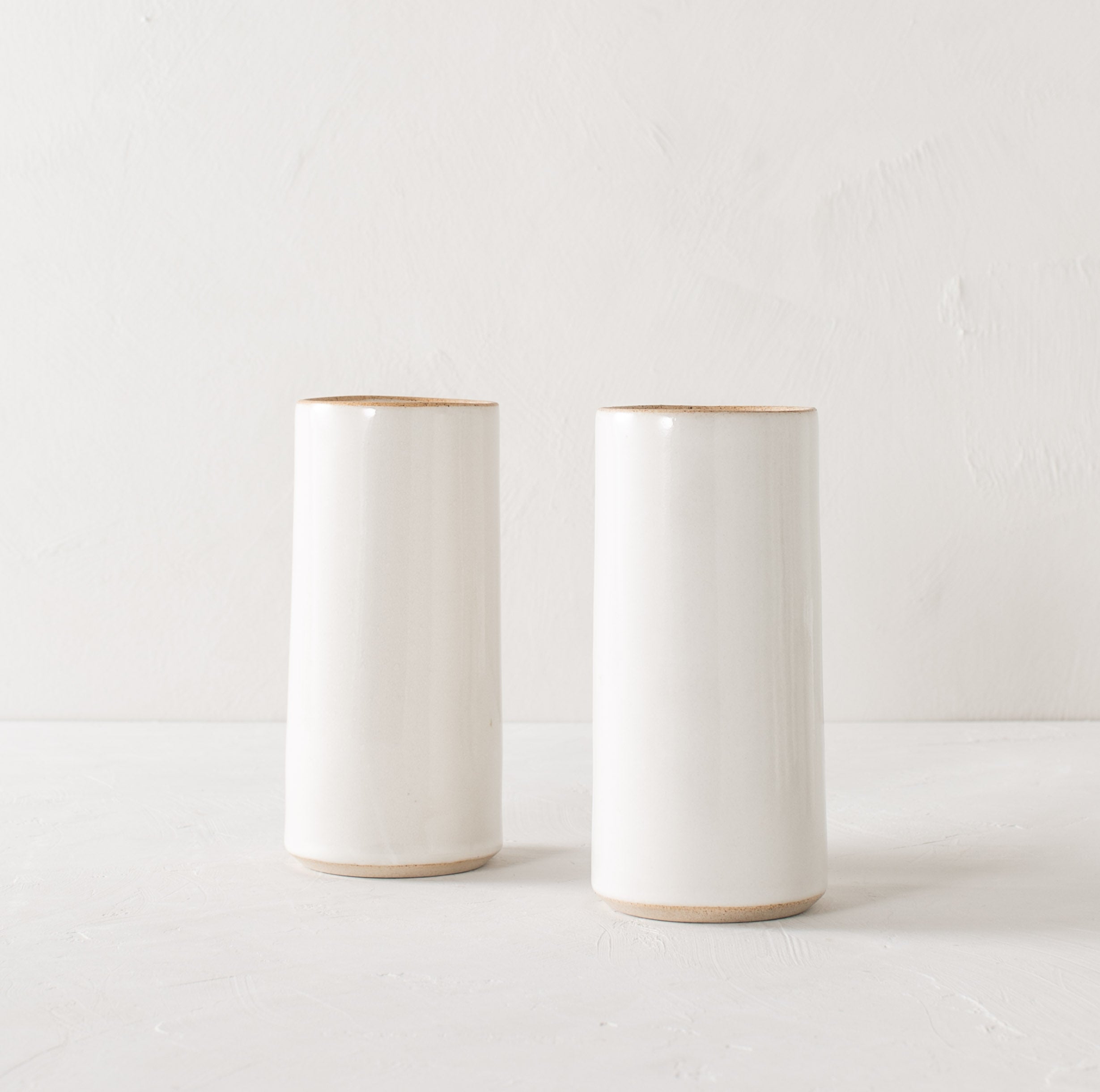 Two tall white glazed ceramic cylinder vases with an exposed stoneware rim and base. Staged on a white textured plaster table and a white plaster textured wall. Designed and sold by Convivial Production, Kansas City Ceramics.