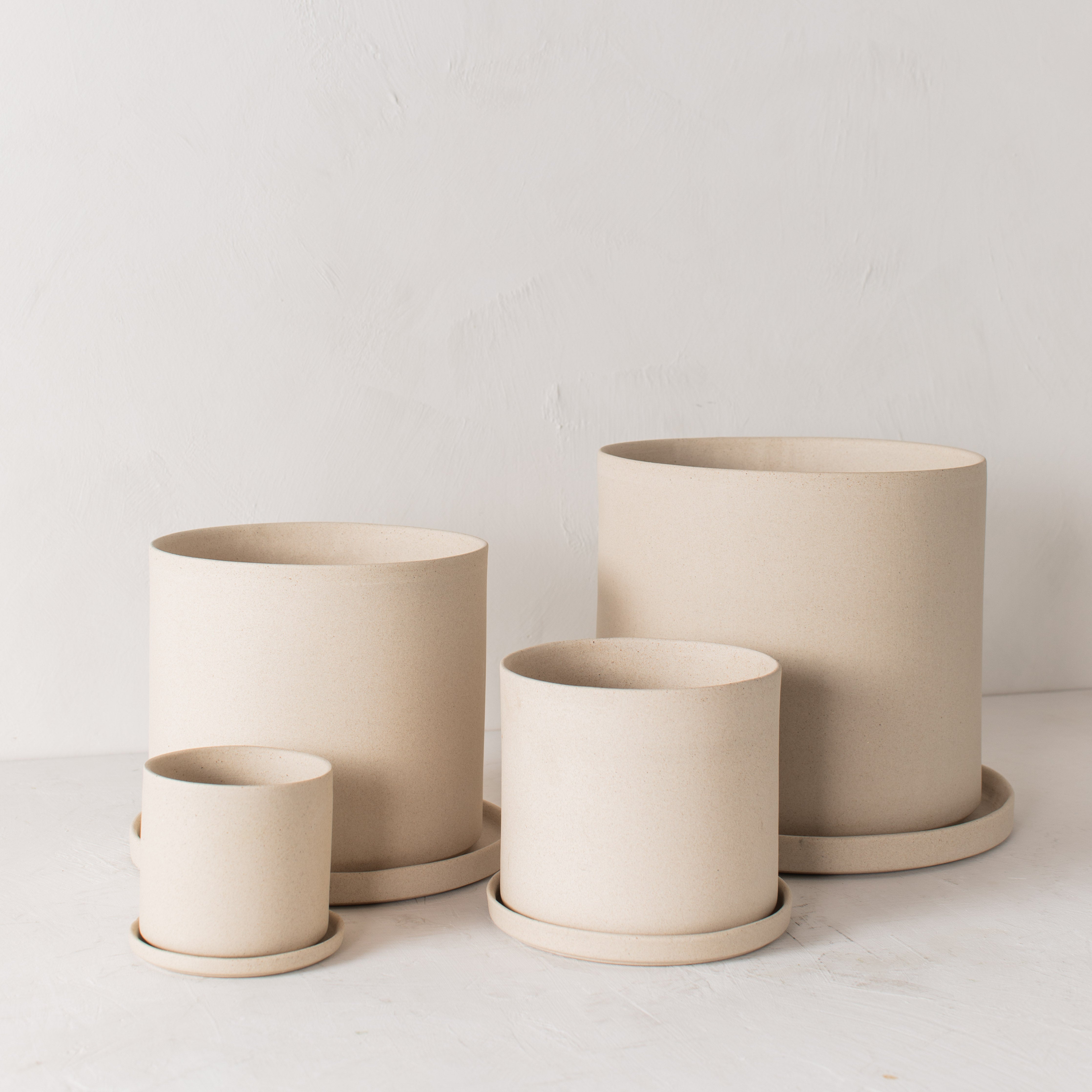 Four stoneware ceramic planters with bottom drainage dishes, 4, 6, 8, and 10 inches. Staged on a white plaster textured tabletop against a plaster textured white wall. Large ta. Designed and sold by Convivial Production, Kansas City Ceramics.
