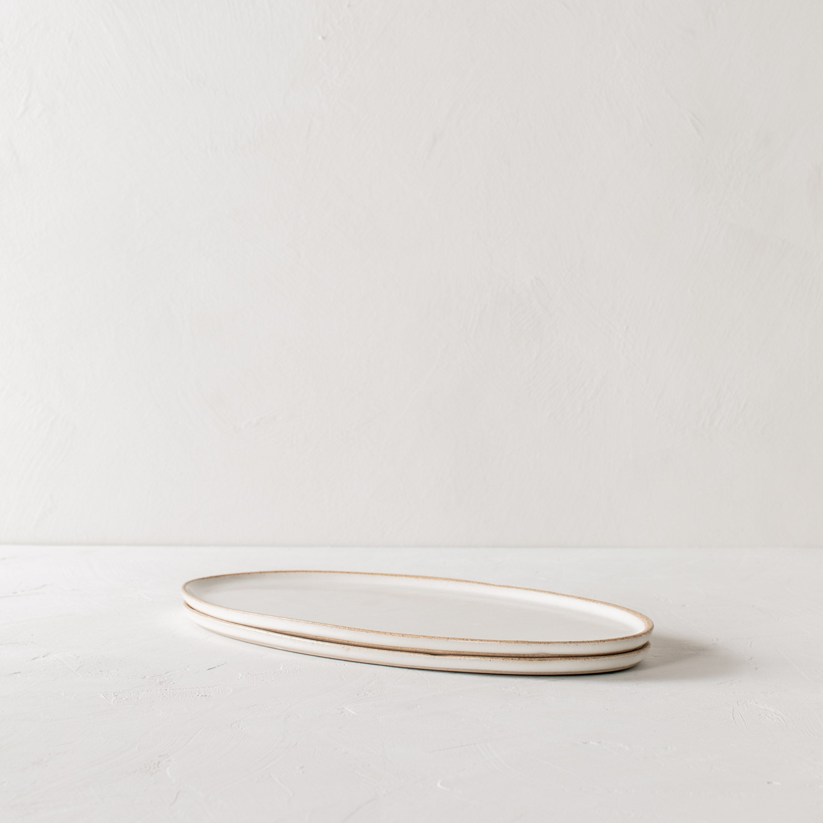 Two narrow vertical ceramic oval serving trays stacked on a white textured table-top. White ivory glazed with exposed stoneware rim. Handmade ceramic serving tray, designed and sold by Convivial Production, Kansas City ceramics.  