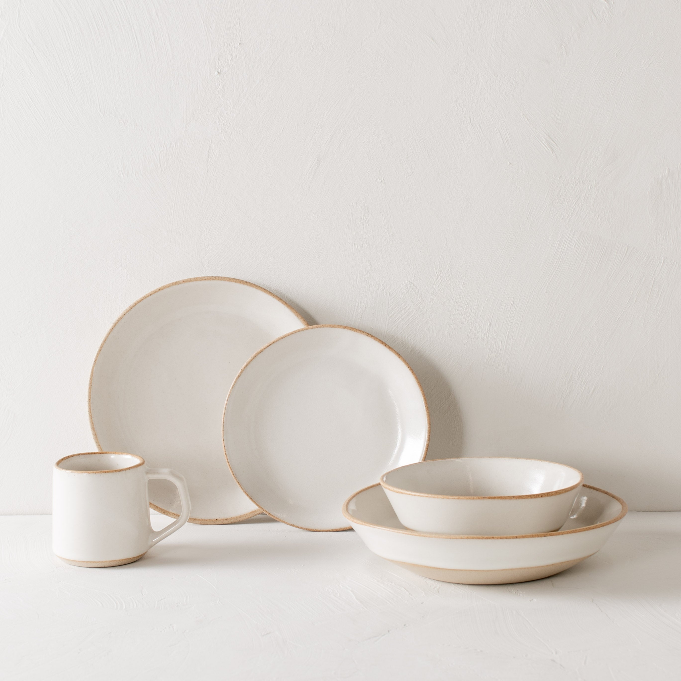 Full ceramic dinnerware set, exposed stoneware rims and bases. All items stylized and stacked overlapping other stacked products. From left to right, minimal mug, dinner and salad plate leaning against white textured wall, minimal bowl nested in pasta bowl. Handmade ceramic dining set designed and sold by Convivial Production, Kansas City ceramics.