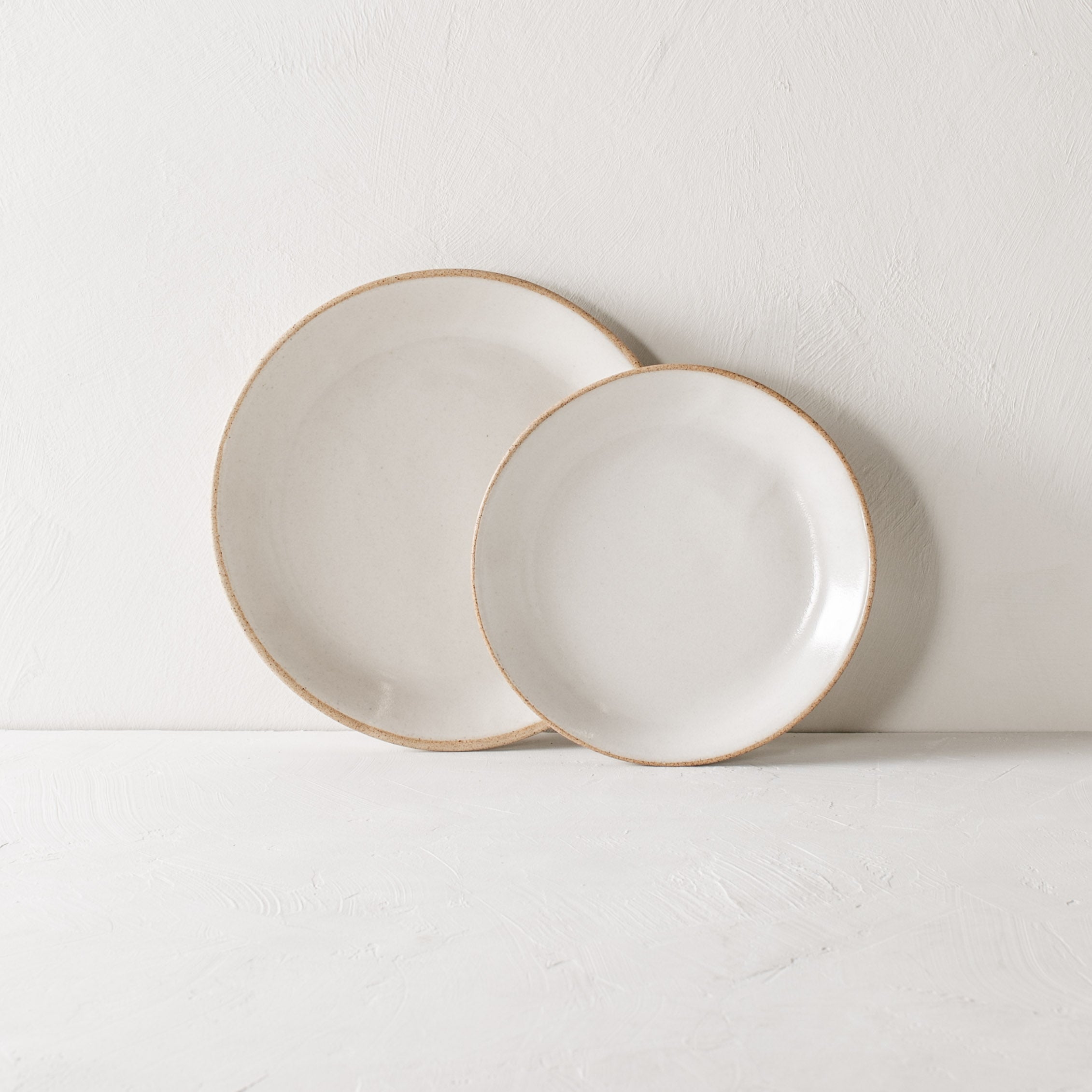 Minimal white ceramic plates against a lime wash wall and white textured tabletop. Plates are different sizes, from left to right: dinner plate, white glaze with an exposed stoneware rime, over lapping the dinner plate is a minimal salad plate identical in look but smaller. Handmade ceramic plates designed and sold by Convivial Production. Kansas City ceramics.