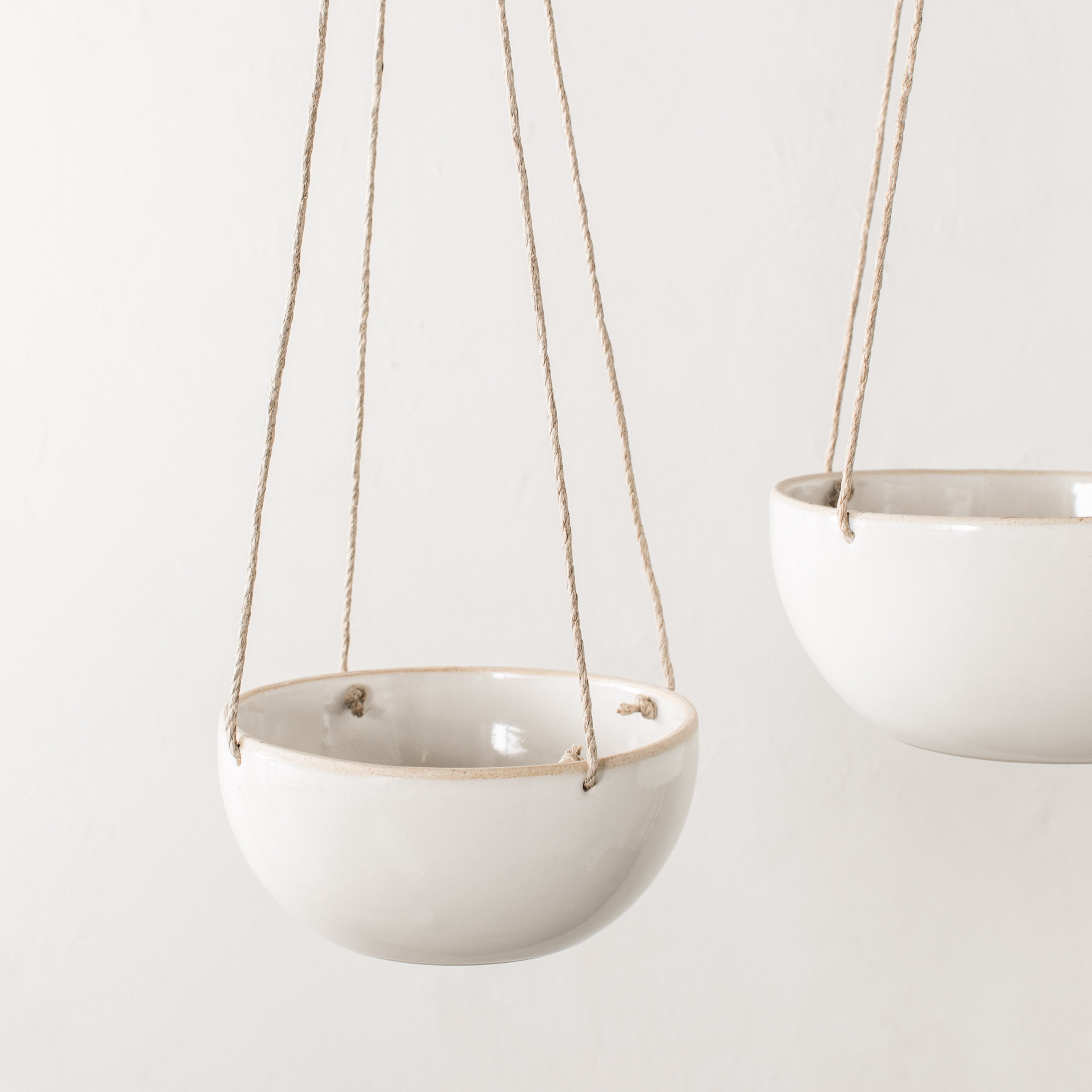 Two minimal ceramic hanging planter. From left to right, 4 inch plater with a plant hangs by a tan hemp cord next to a hanging 6 inch planter. Close up image. Handmade ceramic hanging planter, designed and sold by Convivial Production, Kansas City ceramics. 