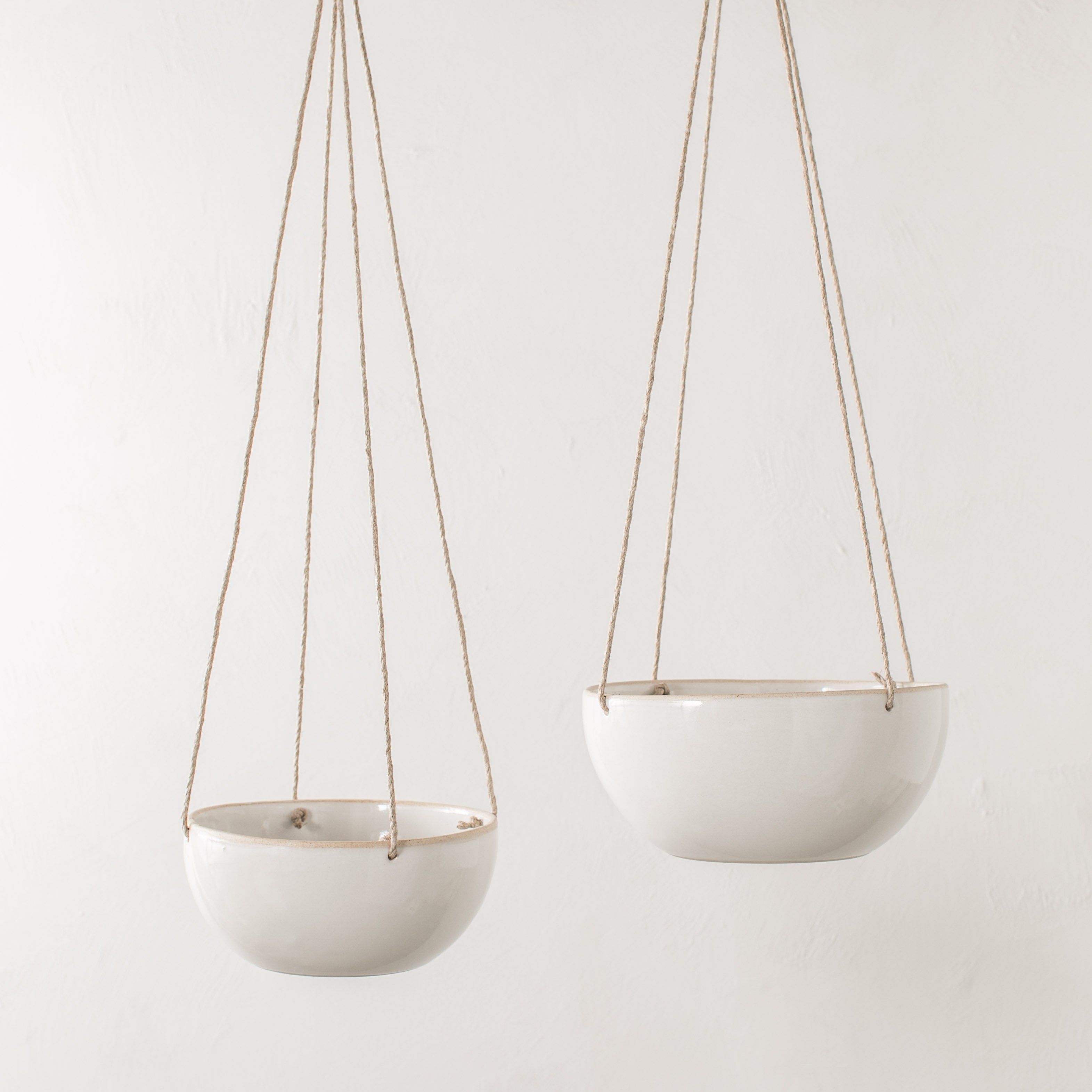 Two minimal ceramic hanging planter. Planters have an exposed stoneware rim and base. From left to right, 4 inch plater with a plant hangs by a tan hemp cord next to a hanging 6 inch planter with a plant. Handmade ceramic hanging planter, designed and sold by Convivial Production, Kansas City ceramics.