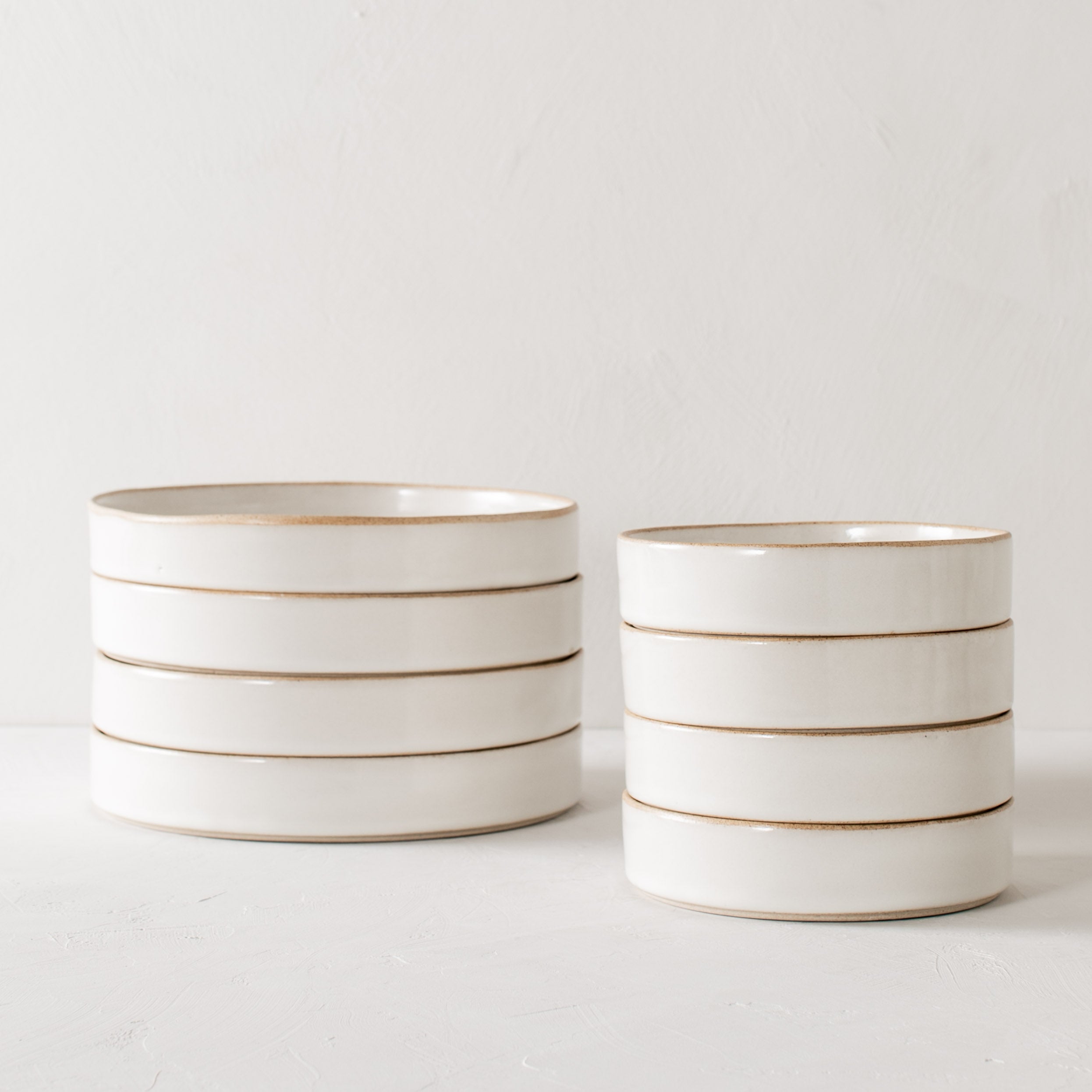 Two stacks of minimal dishes. From left to right, stack of four dinner dishes, stack of salad dishes. Dishes have tall walls with exposed stoneware rims and bases. Handmade ceramic dishes designed and sold by Convivial Production, Kansas City ceramics.