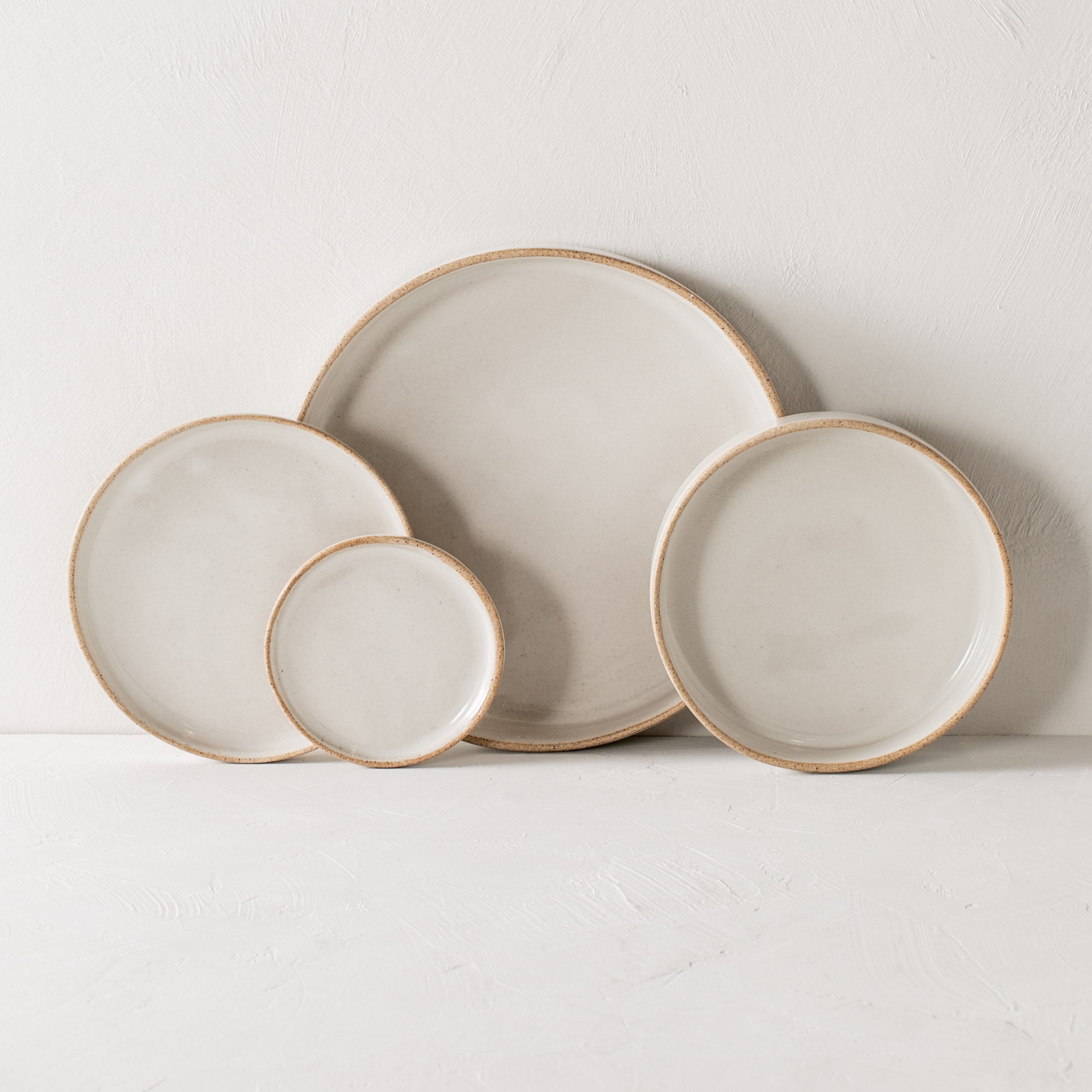 Dish set, leaning against lime wash white backdrop wall and white textured tabletop. From left to right; minimal side 6 inch dish, 4 inch side dish, dinner dish, salad dish. Dishes have a lifted edges creating a deep dish, ceramic white glaze with exposed stoneware rims. Handmade ceramic deep dishes designed and sold by Convivial Production, Kansas City ceramics.  