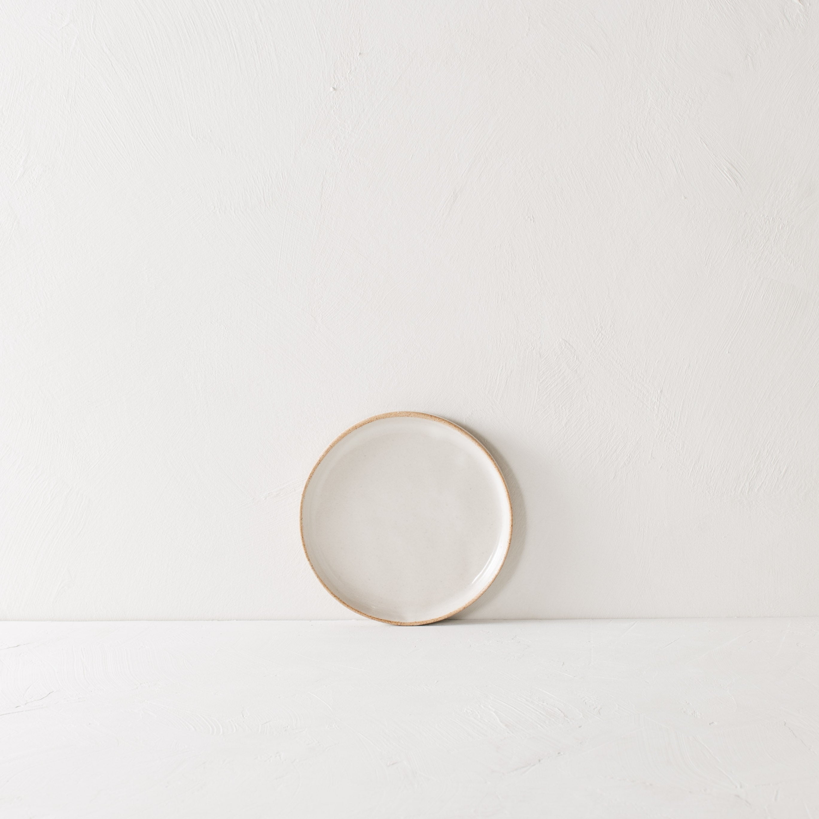 Minimal white 6 inch side dish leaning against the backdrop. Exposed stoneware rim can be seen. Handmade ceramic 6 inch side dish, designed and sold by Convivial Production, Kansas City ceramics.