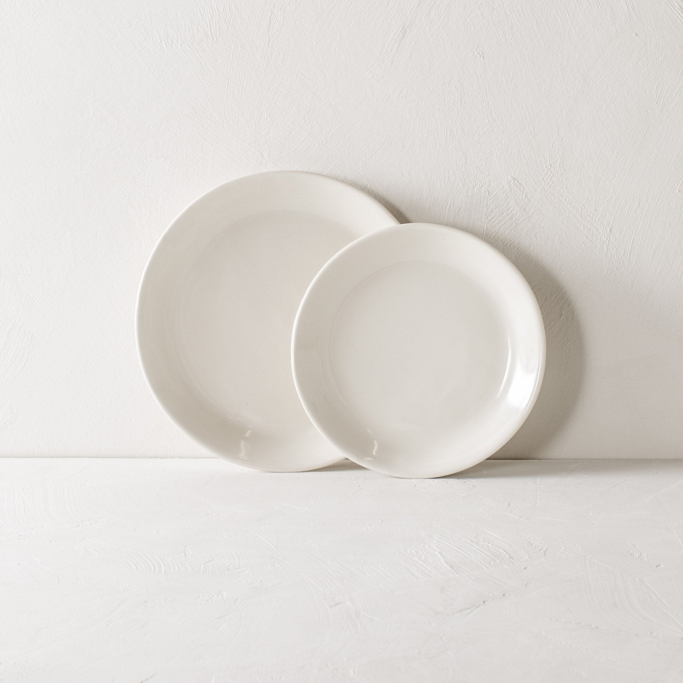 Two minimal white plates, plates are leaning against the textured wall, dinner plate layered under the salad plate. Handmade ceramics plate designed and sold by Convivial Production, Kansas City ceramics.