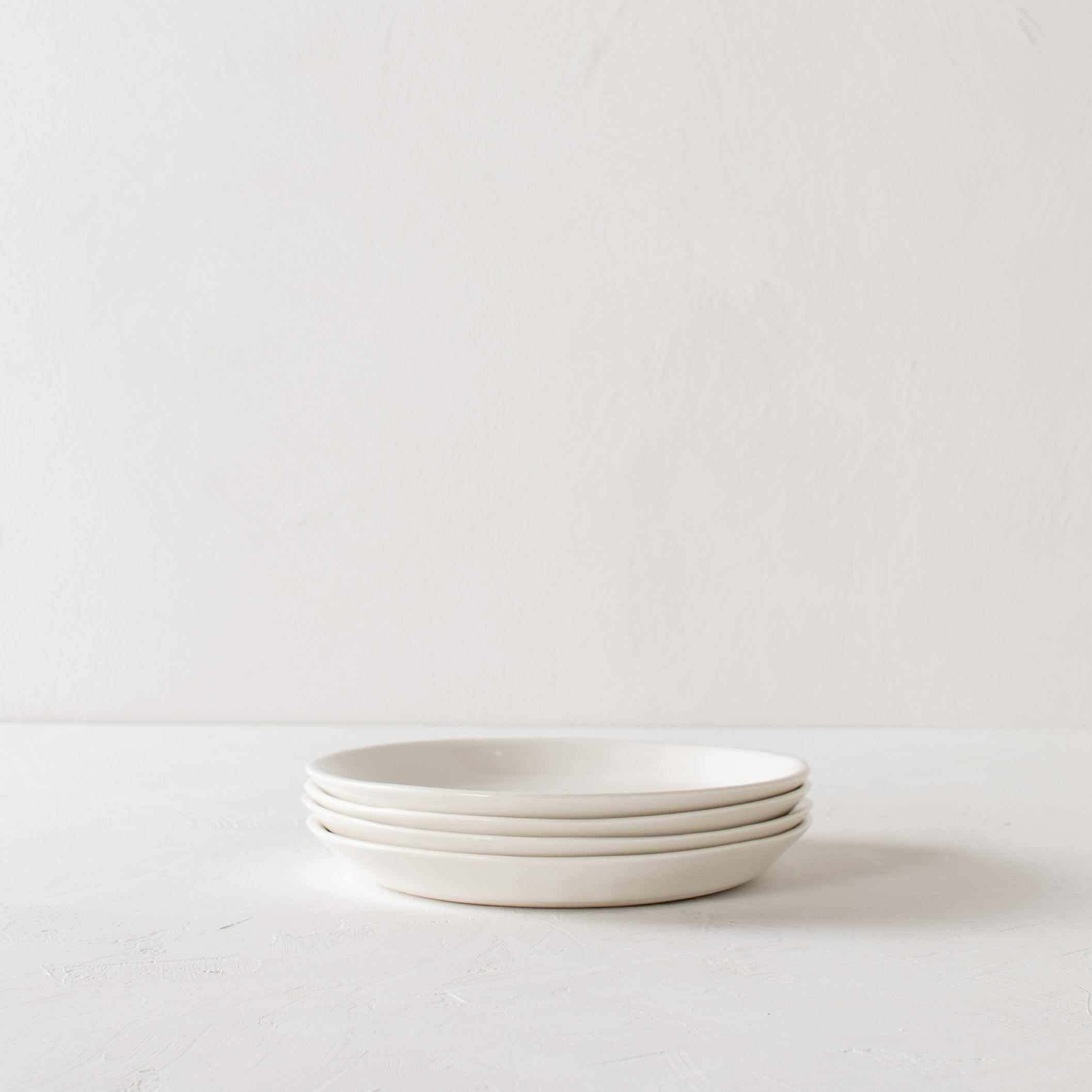 Stacked minimal salad plates. Four stacked plates on a textured white tabletop and white textured backdrop. Handmade ceramic plates designed and sold by Convivial Production, Kansas City Ceramics.