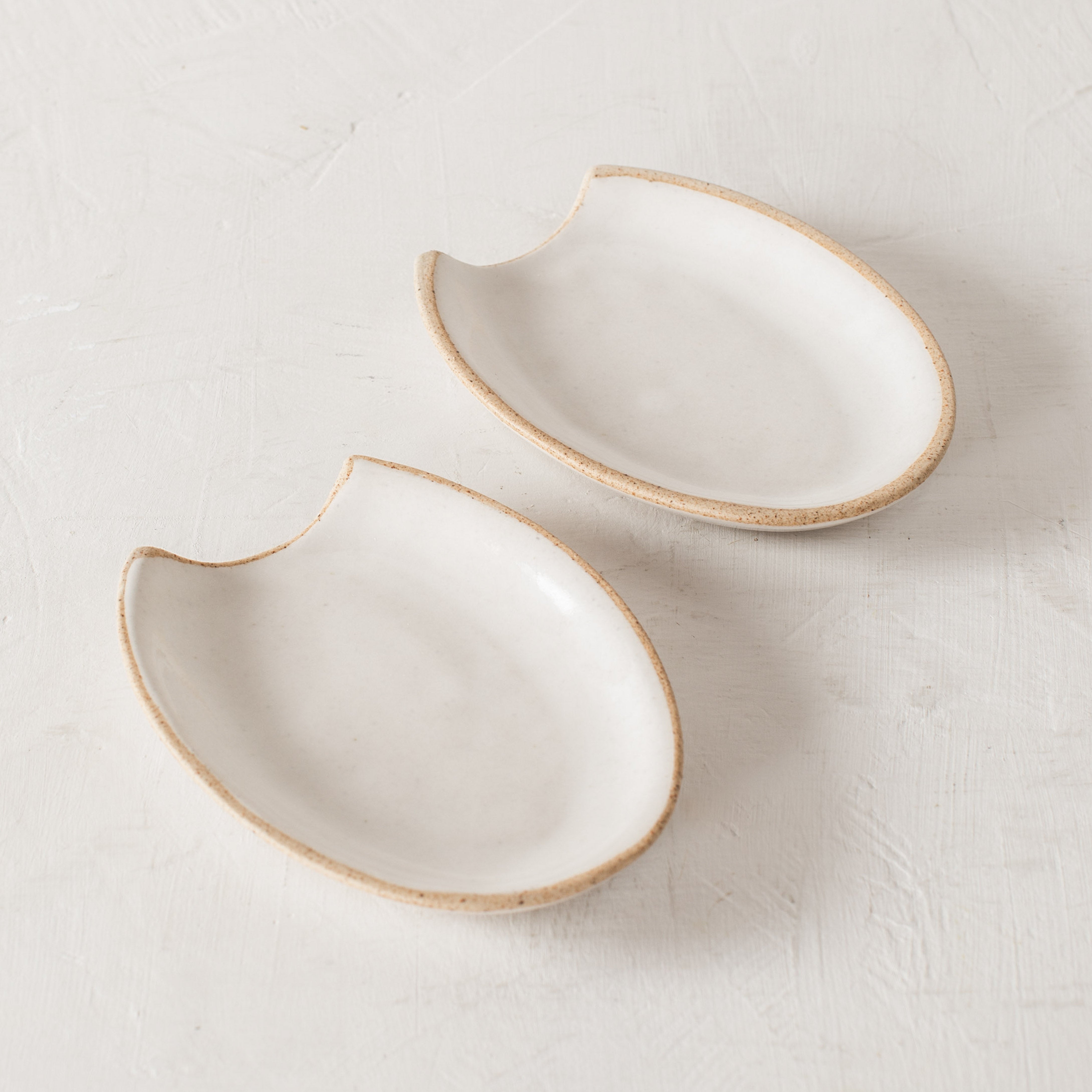 Two arch shaped ceramic spoon rests side by side. Image from above, white minimal arched shaped with an exposed rim. Handmade ceramic spoon rest designed and sold by Convivial Production, Kansas City ceramics.