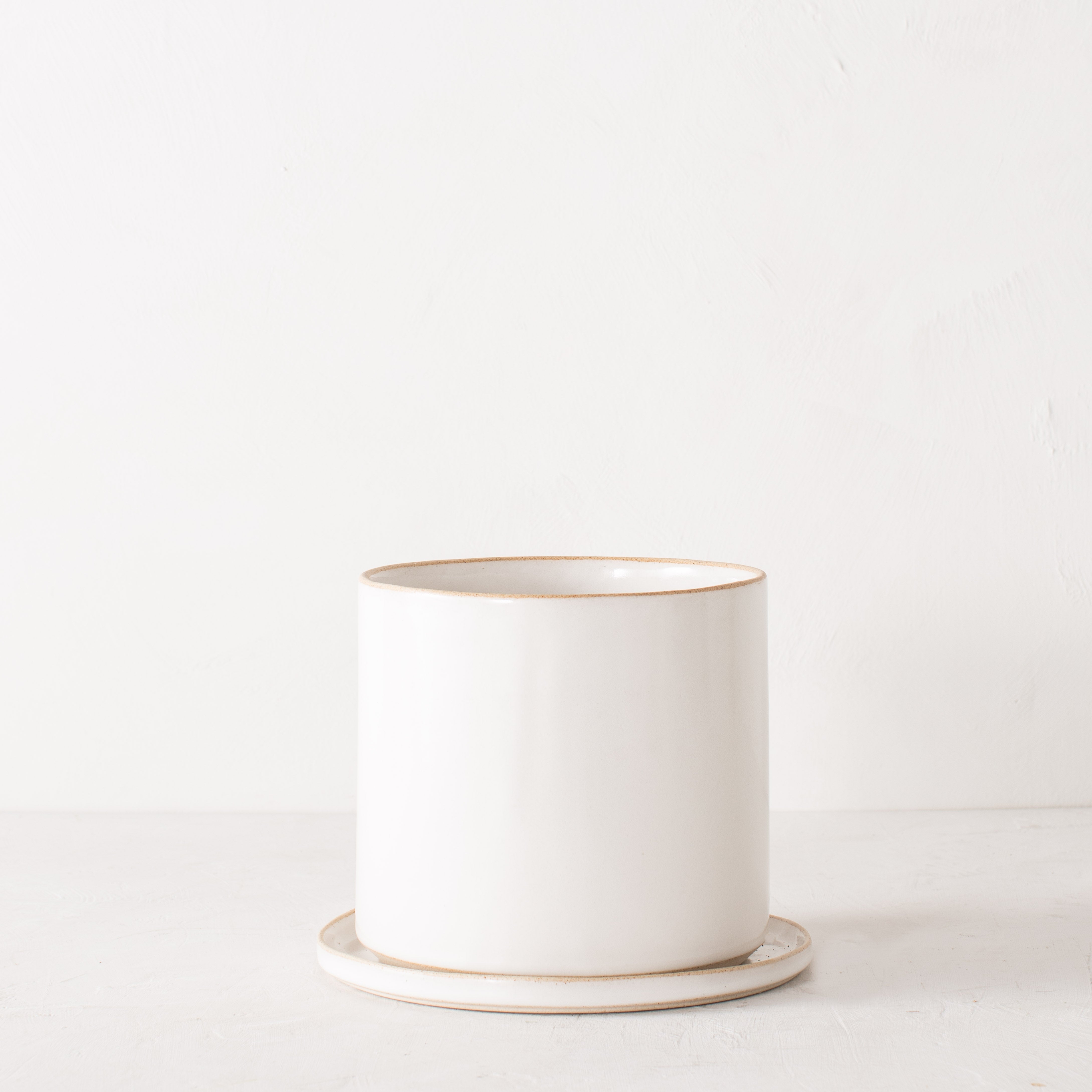8 inch white ceramic planter with a bottom drainage dishes. Staged on a white plaster textured tabletop against a plaster textured white wall. Designed and sold by Convivial Production, Kansas City Ceramics.