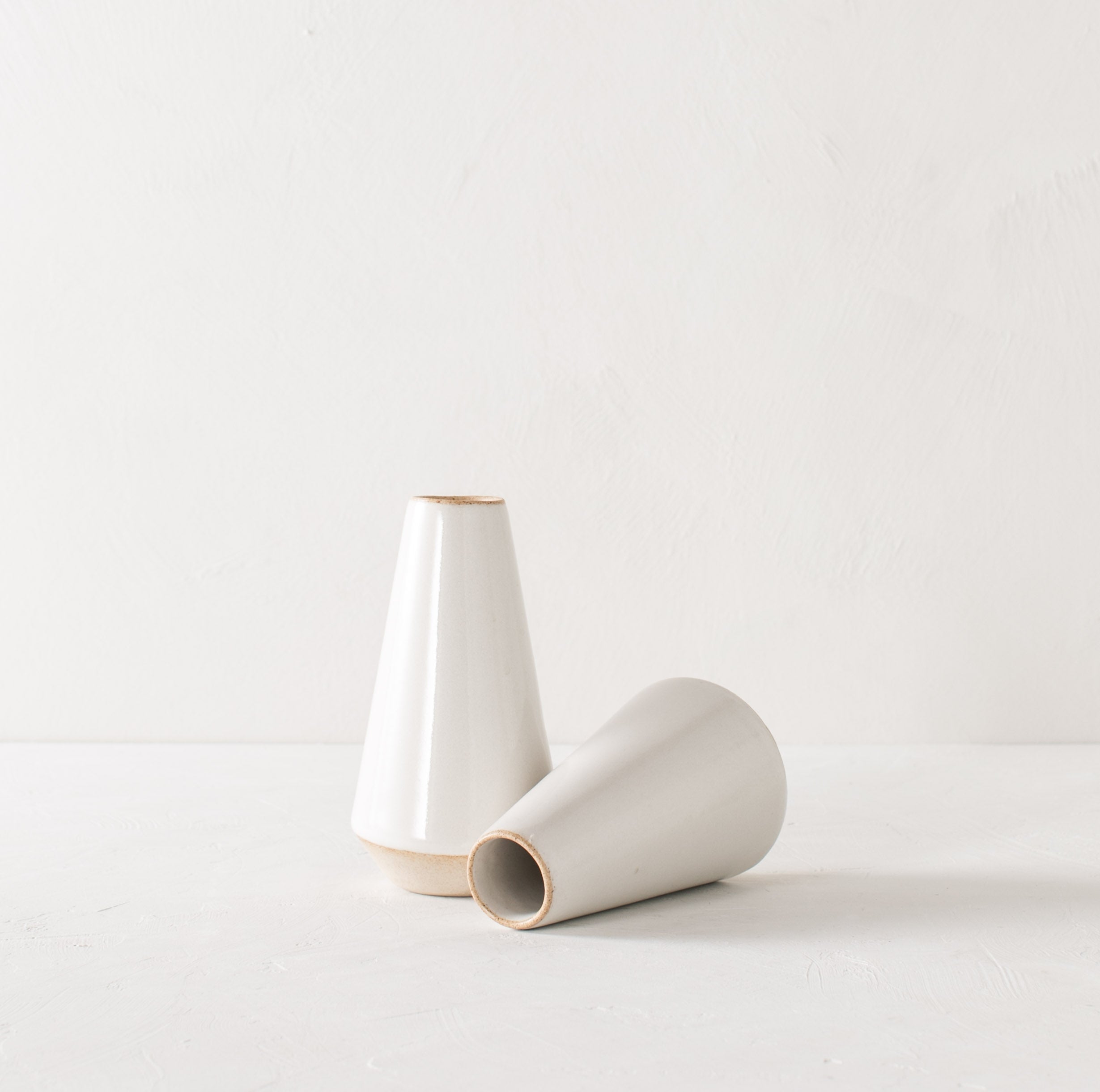 Two minimal white 7 inch tapered bud vases. White glazed with an exposes stoneware rim and base. One upright while the other lays on its side. Handmade ceramic bud vase designed and sold by Convivial Production, Kansas City Ceramics.
