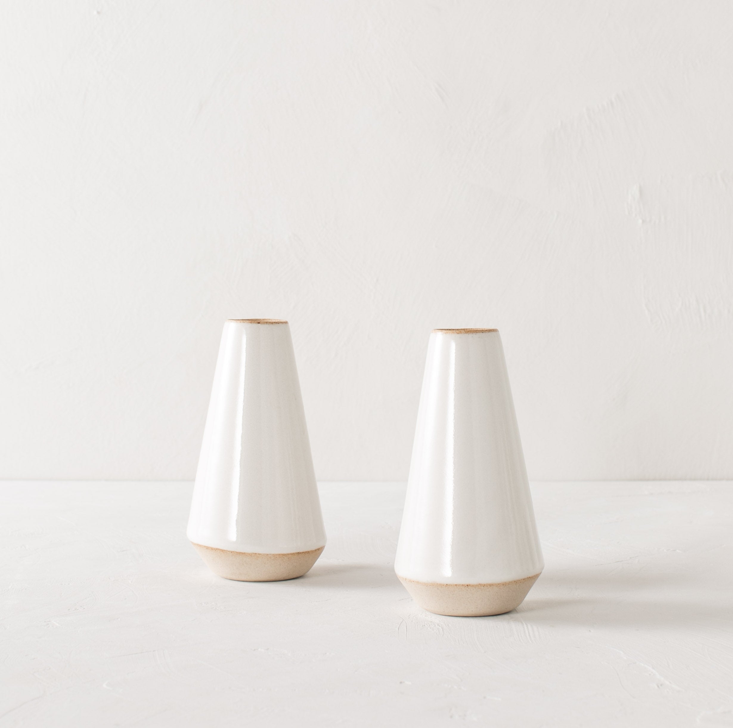 Two minimal white 7 inch tapered bud vase. White glazed with an exposes stoneware rim and base. Both upright side by side on a white plaster textured tabletop and white textured back drop. Handmade ceramic bud vase designed and sold by Convivial Production, Kansas City Ceramics.