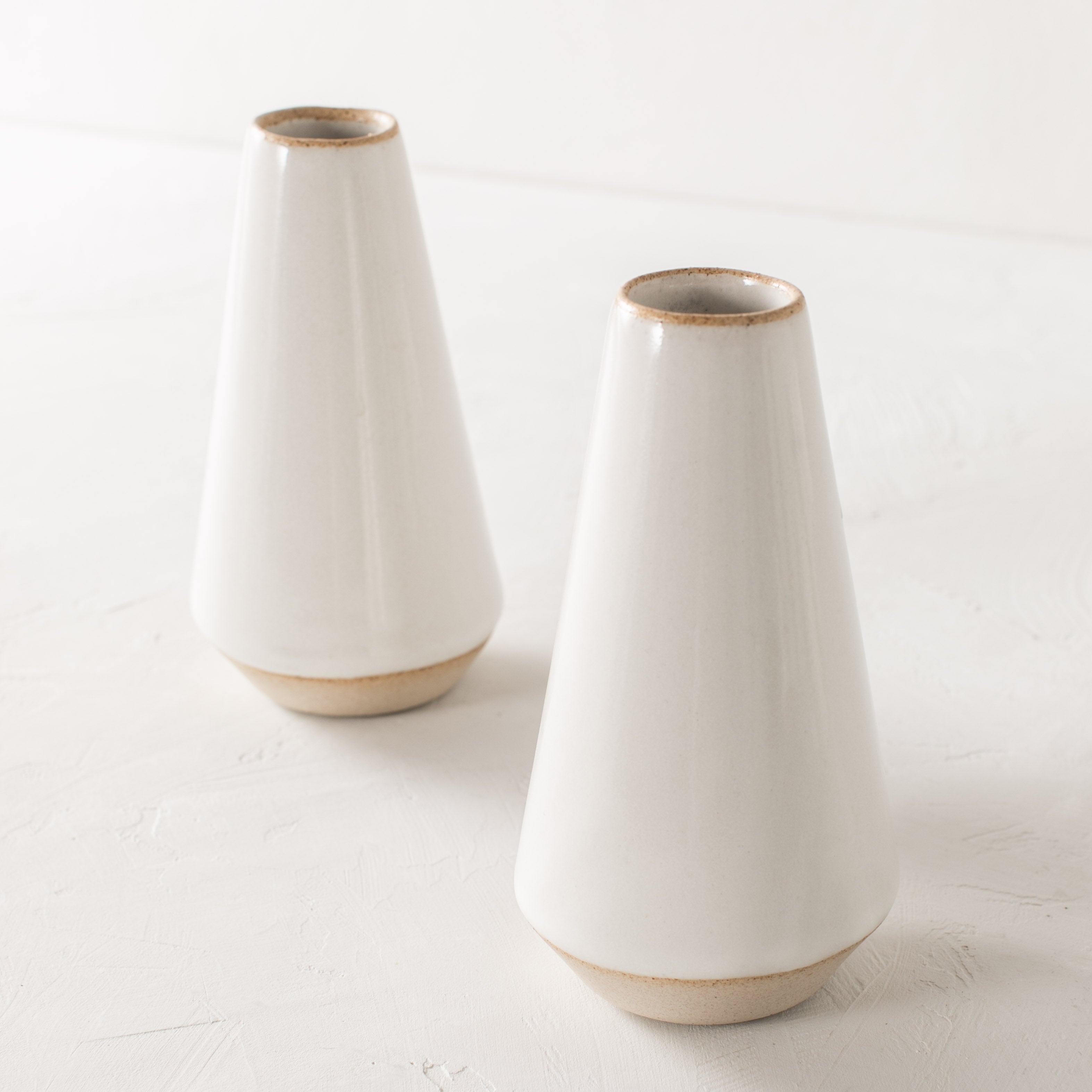 Two minimal white 7 inch tapered bud vase. White glazed with an exposes stoneware rim and base. Both upright side by side on a white plaster textured tabletop and white textured back drop. Handmade ceramic bud vase designed and sold by Convivial Production, Kansas City Ceramics.