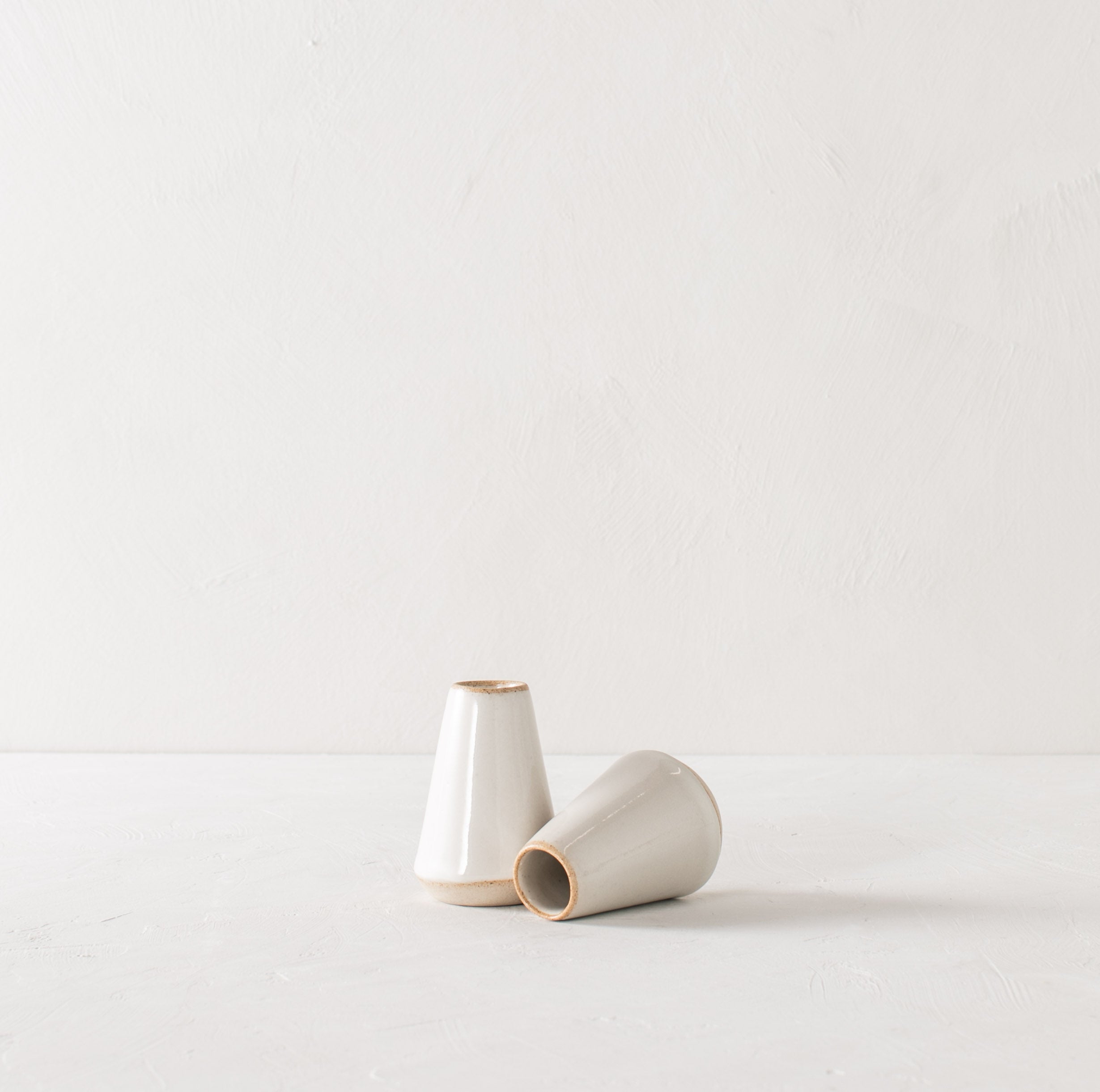 Two minimal white 4 inch tapered bud vase. White glazed with an exposes stoneware rim and base. One upright while the other lays on its side. Handmade ceramic bud vase designed and sold by Convivial Production, Kansas City Ceramics.