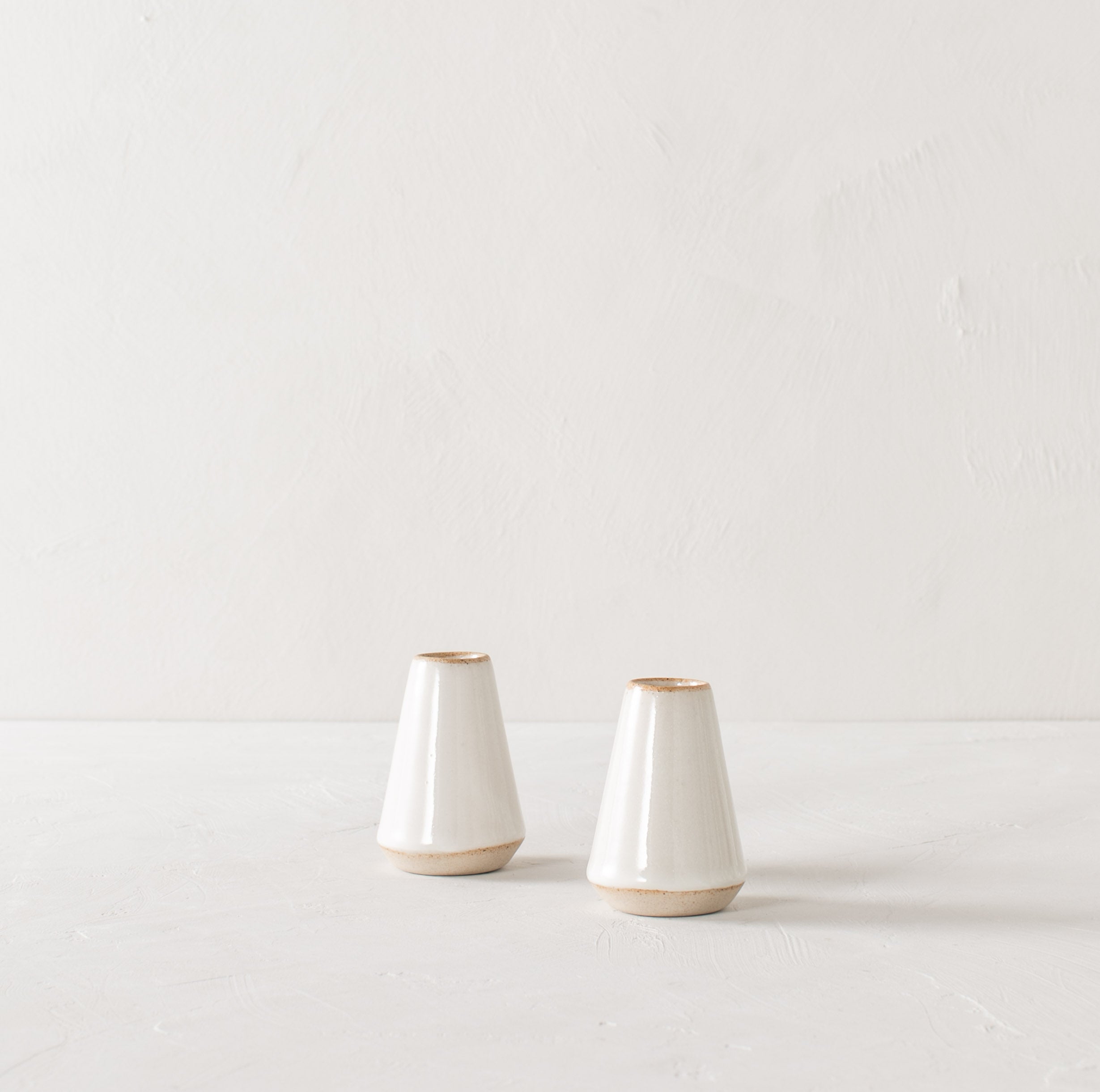 Two minimal white 4 inch tapered bud vase. White glazed with an exposes stoneware rim and base. Both upright side by side on a white plaster textured tabletop and white textured back drop. Handmade ceramic bud vase designed and sold by Convivial Production, Kansas City Ceramics.