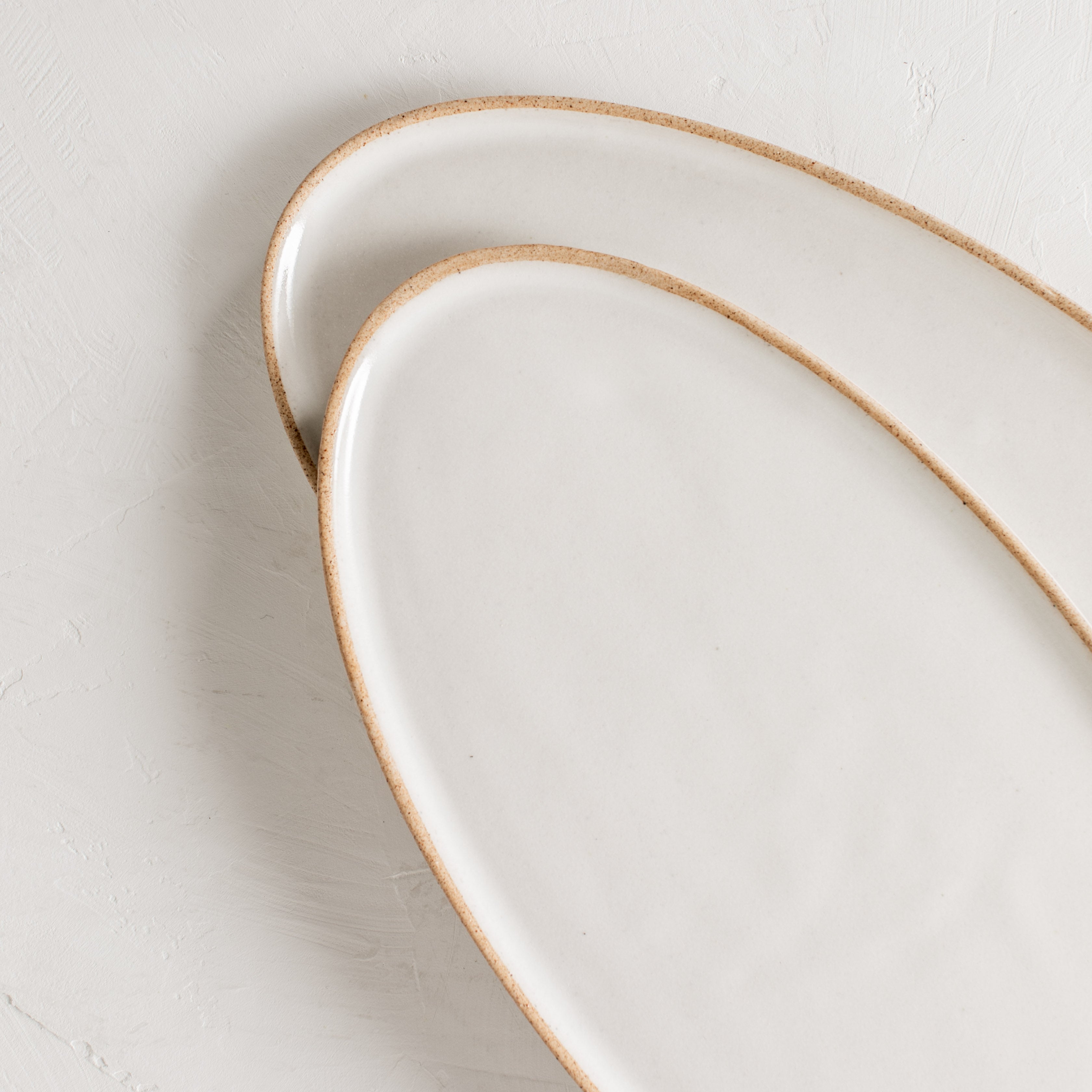 Two narrow vertical ceramic oval serving trays staggered and overlapping on a white textured table-top. Close up of the white ivory glazed with exposed stoneware rim. Handmade ceramic serving tray, designed and sold by Convivial Production, Kansas City ceramics.  