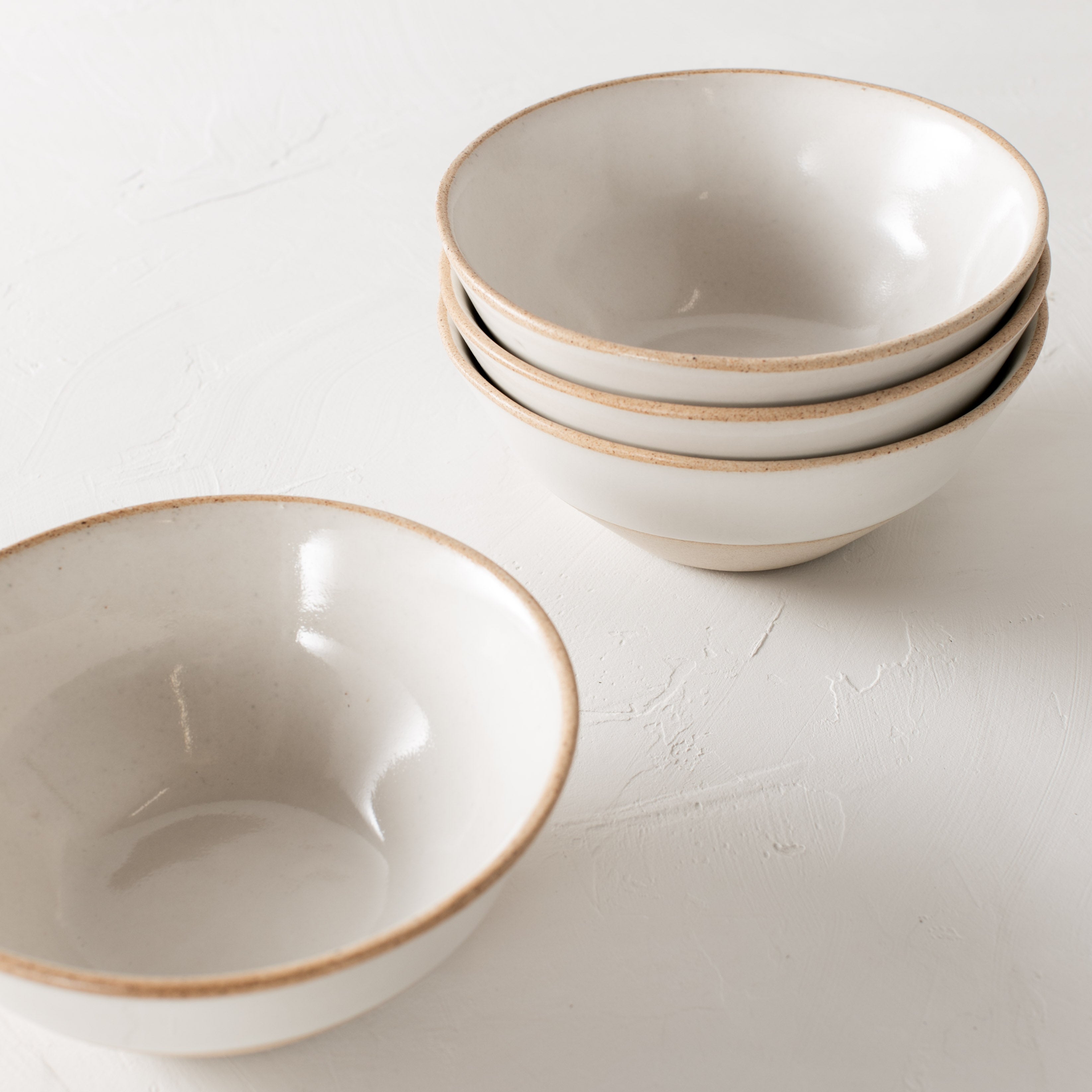 White ceramic bowls. Handmade glazed stoneware bowls, staged together staggered in a group of four. Three stacked and one laid out beside the stack.Bowls have exposed stoneware rims as well as bases. Centered in the image staged against a white textured wall and table top. Designed and sold by Convivial Production, Kansas City Ceramics.