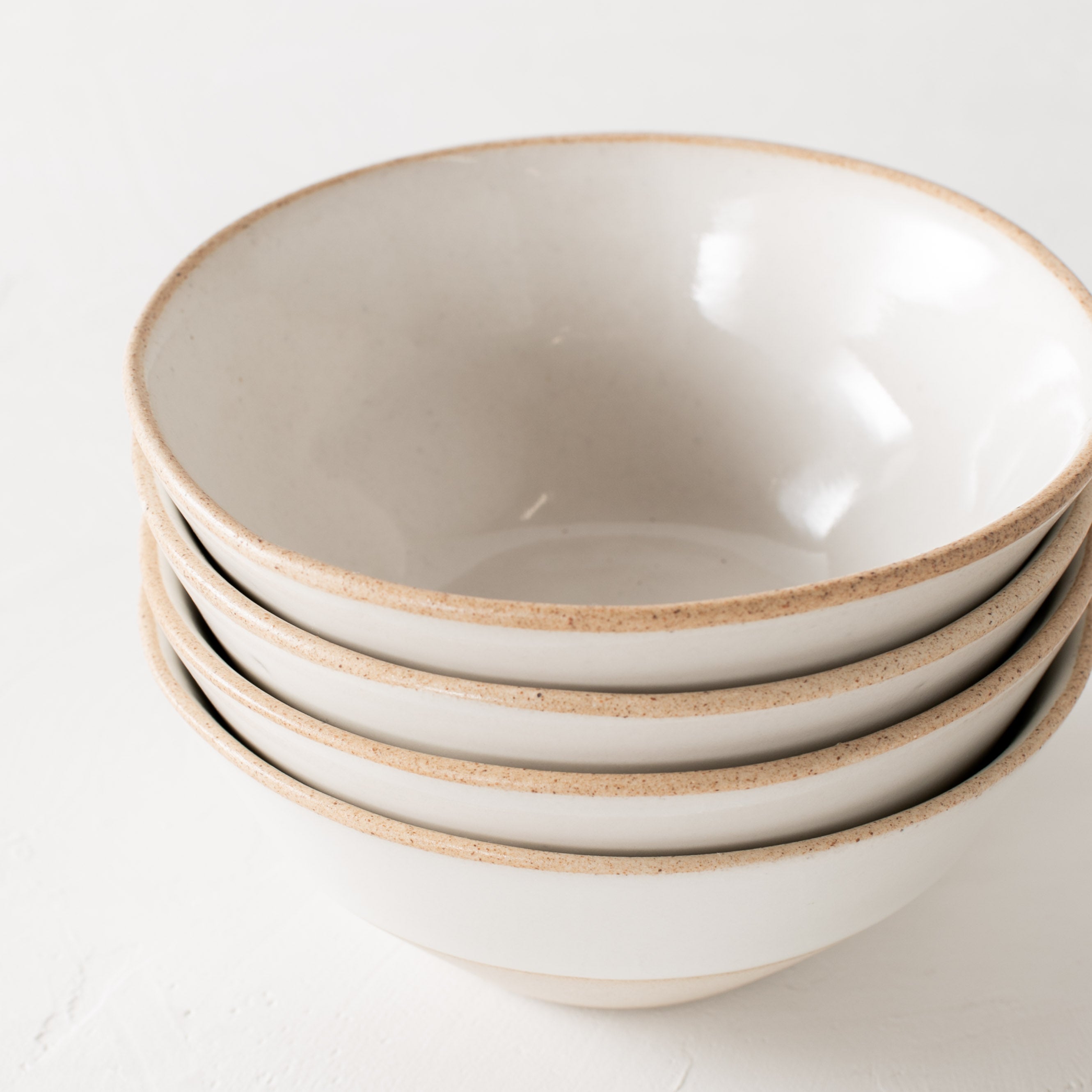 White ceramic bowls. Handmade glazed stoneware bowls, close up of stacked together in a group of four. Bowls have exposed stoneware rims as well as bases. Centered in the image staged against a white textured wall and table top. Designed and sold by Convivial Production, Kansas City Ceramics.