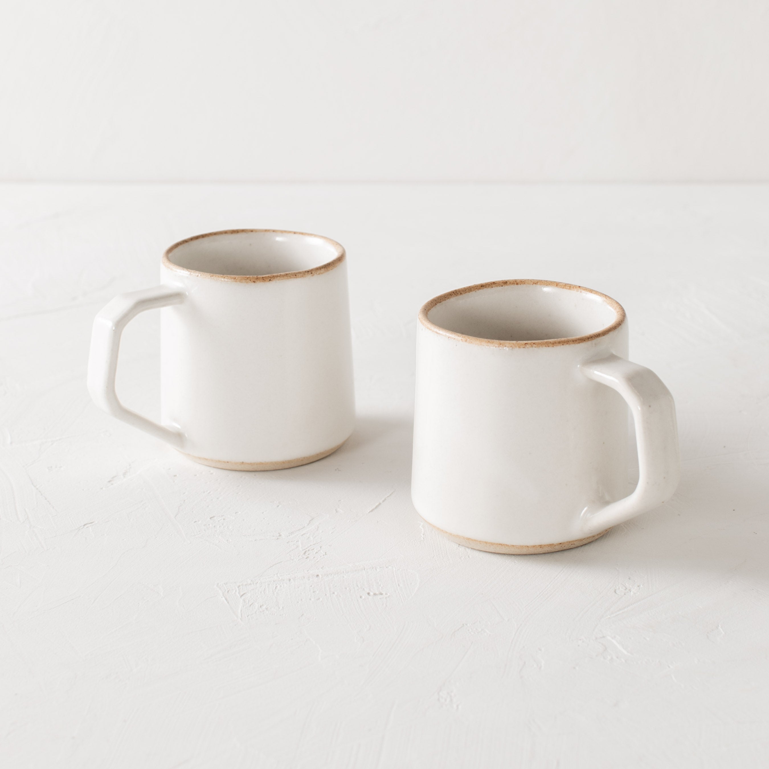 Pair of white minimal ceramic mug is staged on a white textured table top and white textured back drop. Mugs have exposed warm stoneware on the rims as well as the base. Handles are geometric in shape but still rounded. Convivial Production, Kansas City Ceramics. Handmade ceramic mug.