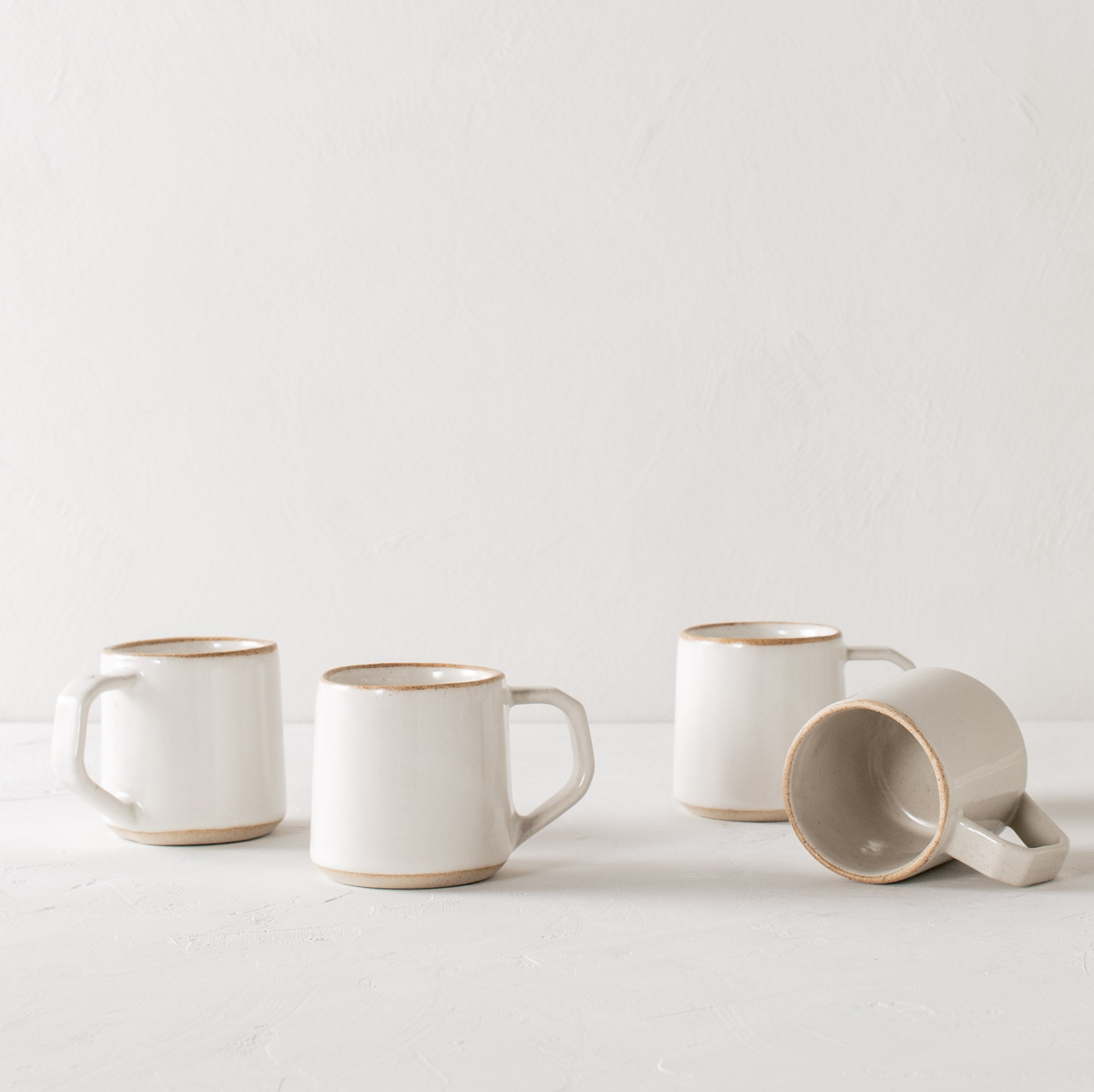 Four white minimal ceramic mugs is staged on a white textured table top and white textured back drop. Mugs have exposed warm stoneware on the rims as well as the base. Handles are geometric in shape but still rounded. Convivial Production, Kansas City Ceramics. Handmade ceramic mug