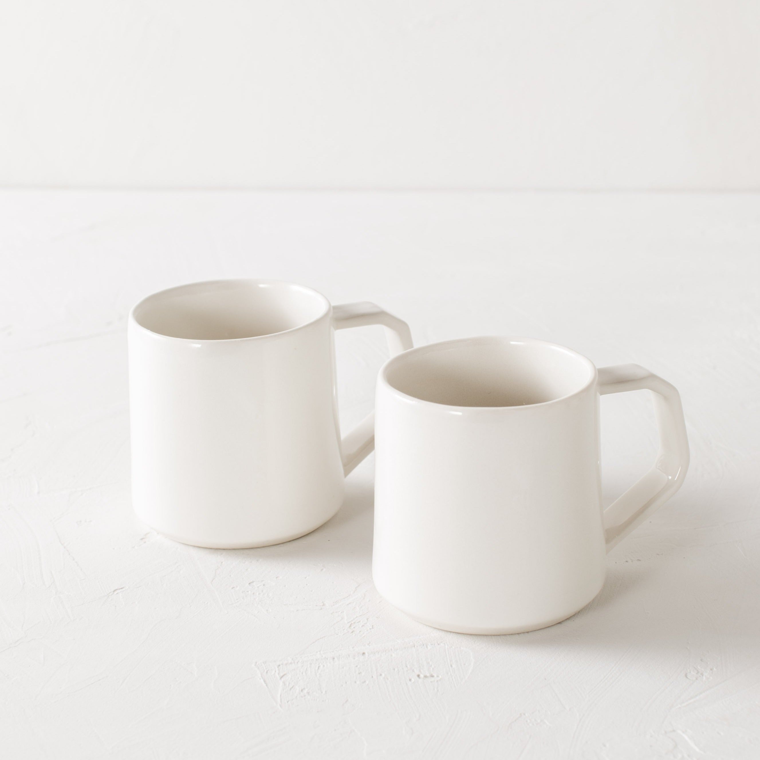 Pair of white minimal ceramic mug is staged on a white textured table top and white textured back drop. Mugs are glazes in ivory white and the handles are geometric in shape but still rounded. Convivial Production, Kansas City Ceramics. Handmade ceramic mug.  