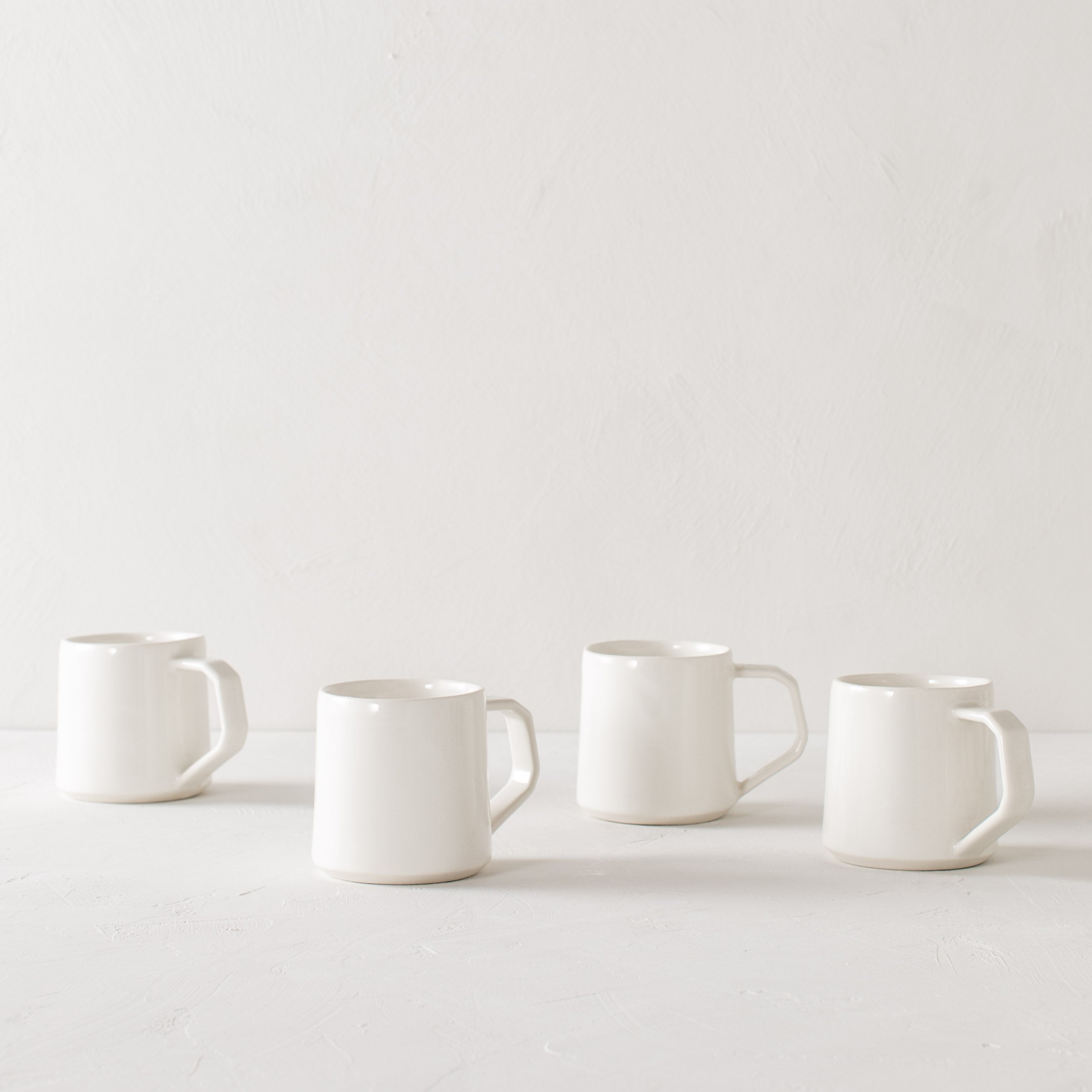 Four white minimal ceramic mugs staged staggered on a white textured table top and white textured back drop. Mugs are ivory white in color and handles are geometric in shape but still rounded. Convivial Production, Kansas City Ceramics. Handmade ceramic mug.  