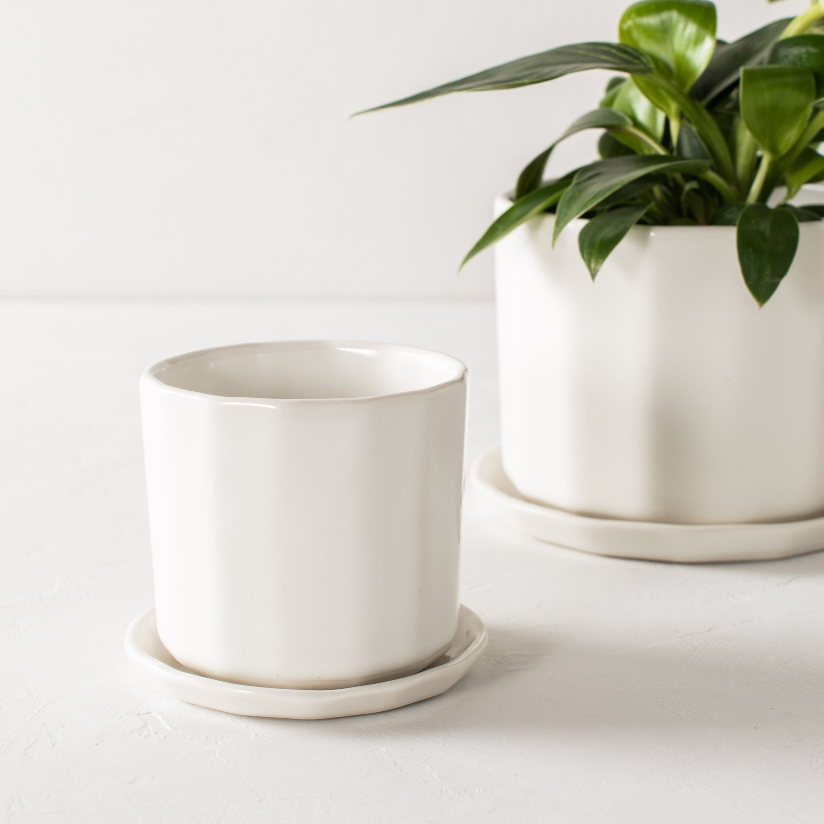 Two white geometric shaped ceramic planters, one empty sitting upright in front of a larger geometric planter with a plant inside also with a bottom drainage dish. Staged on a white plaster textured tabletop against a plaster textured white wall. Designed and sold by Convivial Production, Kansas City Ceramics.