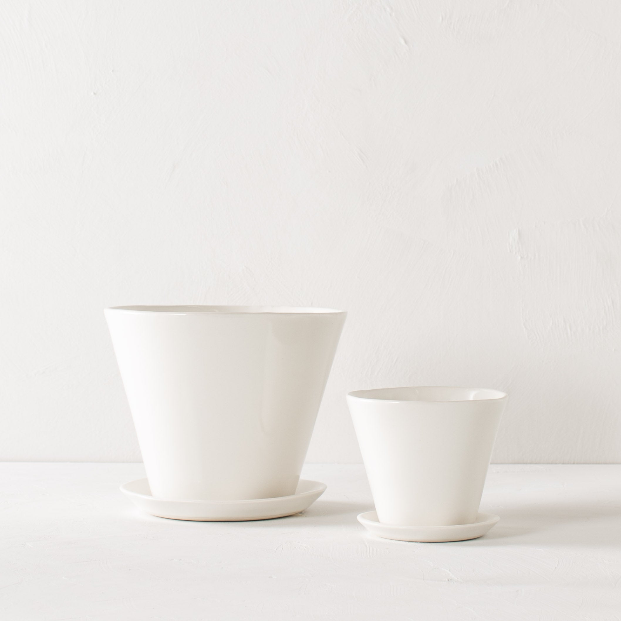 Two white tapered ceramic planter side by side. One larger (7 inches) the other smaller (4 inches ). Both have bottom drainage dishes as well as plants inside. Background is a textured white and the table top mimics the wall design. Designed and sold by Convivial Production, Kansas City Ceramics.