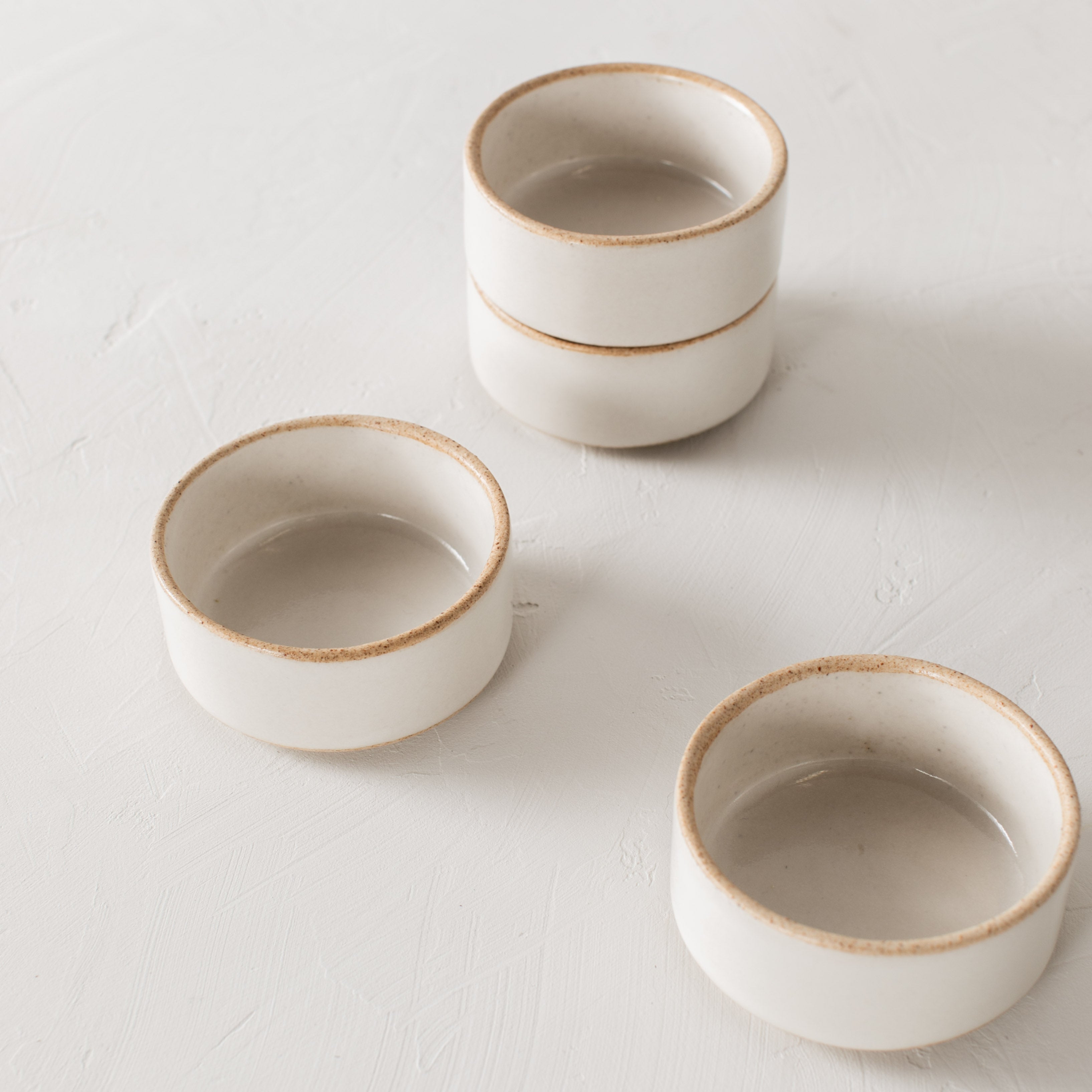 Four ceramic dishes staged on a white tabletop. Two stacked with the other two laid near. Ramekins have an exposed stoneware rim and base. Handmade ceramic ramekin dishes, designed and sold by Convivial Production, Kansas City ceramics.