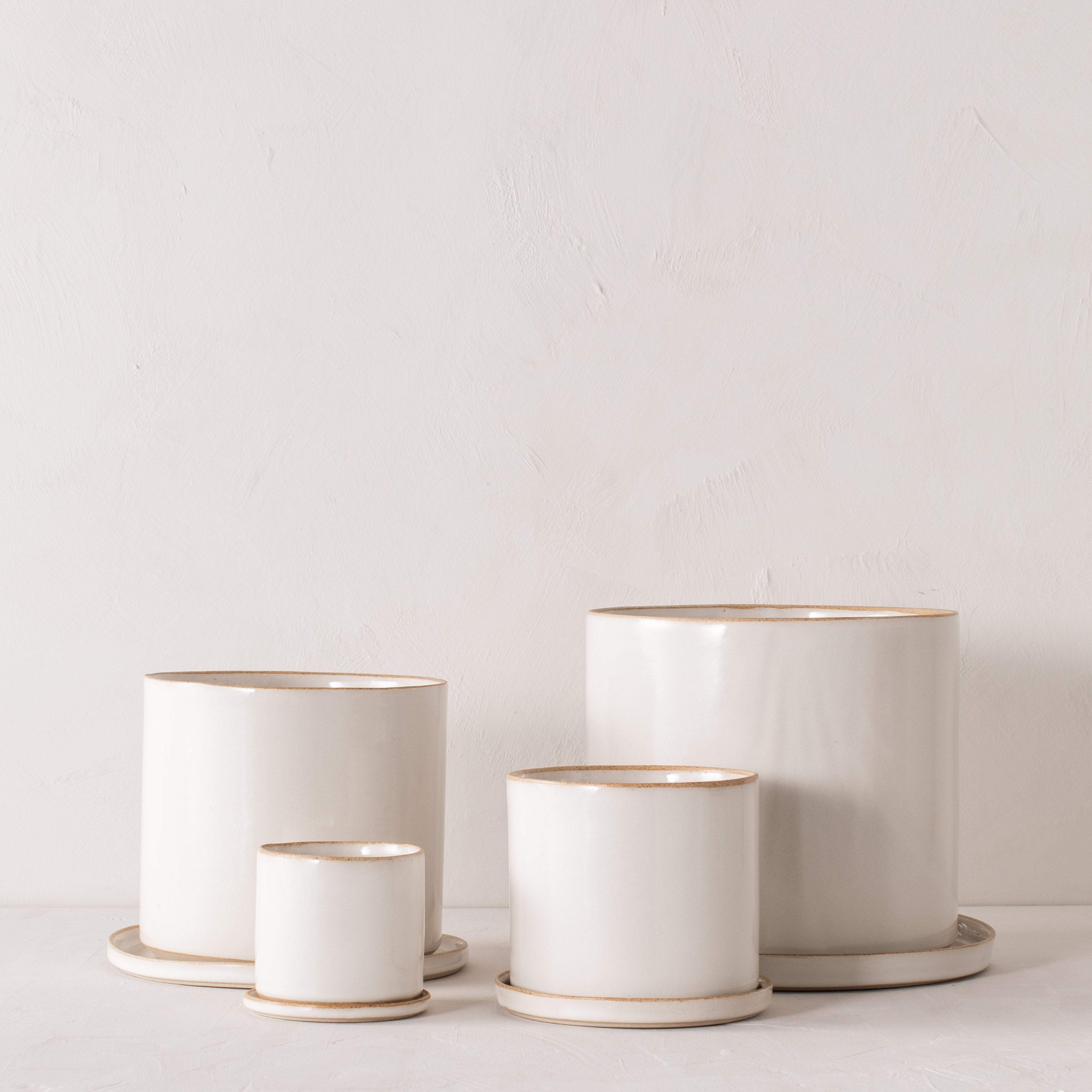 Four white ceramic planters with bottom drainage dishes, 4, 6, 8, and 10 inches. Staged on a white plaster textured tabletop against a plaster textured white wall. Designed and sold by Convivial Production, Kansas City Ceramics.