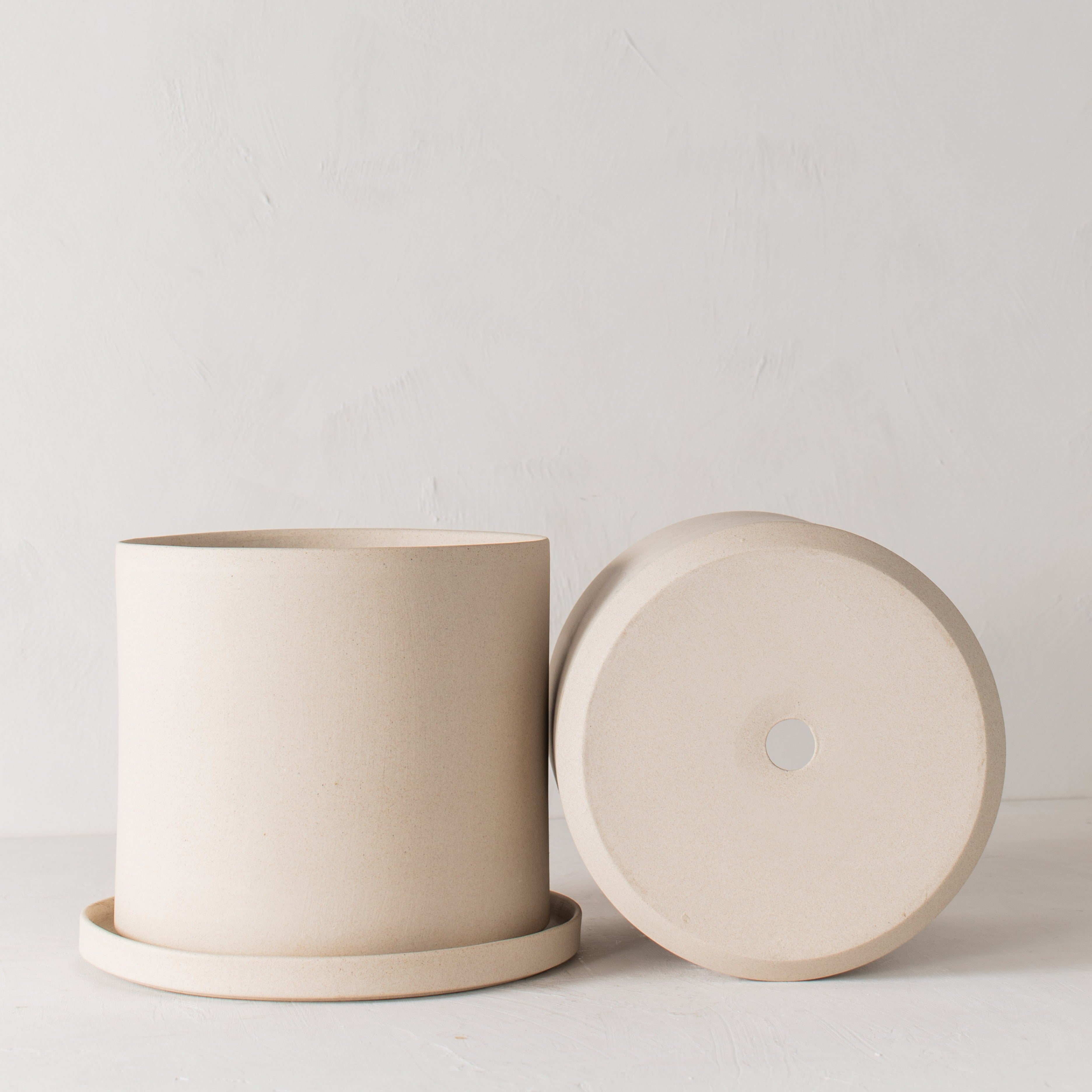 Two stoneware 10 inch ceramic planters with bottom drainage dish, one on its side with a drainage hole. Staged on a white plaster textured tabletop against a plaster textured white wall. Designed and sold by Convivial Production, Kansas City Ceramics.