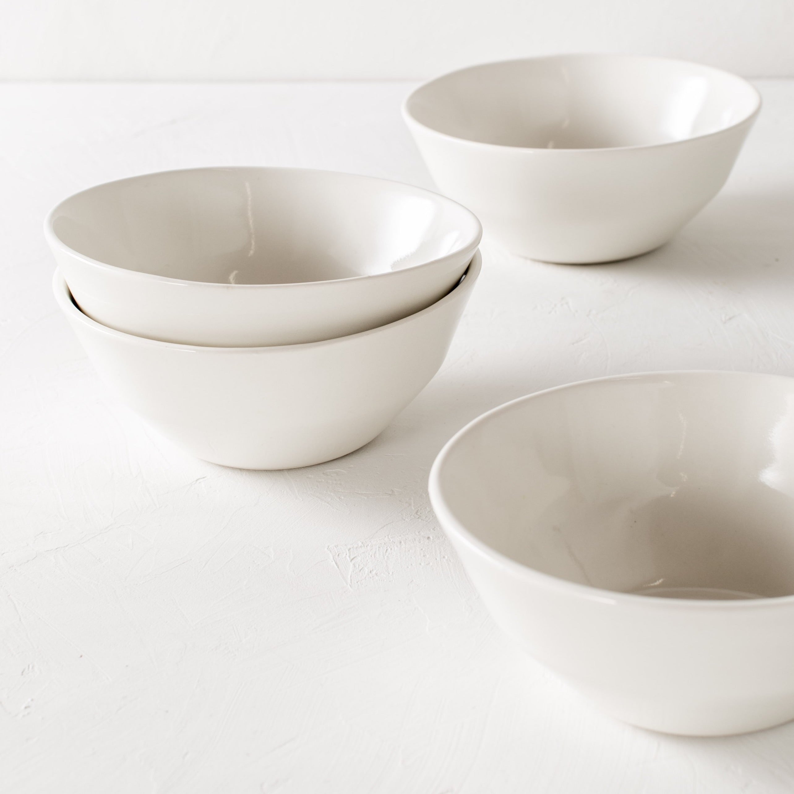 Four white ceramic minimal bowls staged in close up photo. Two are staggered on the table top and the other two are stacked. Staged  on a white textured tabletop against a white textured backdrop. Handmade bowls are designed and sold by Convivial Production. Kansas City Ceramics.
