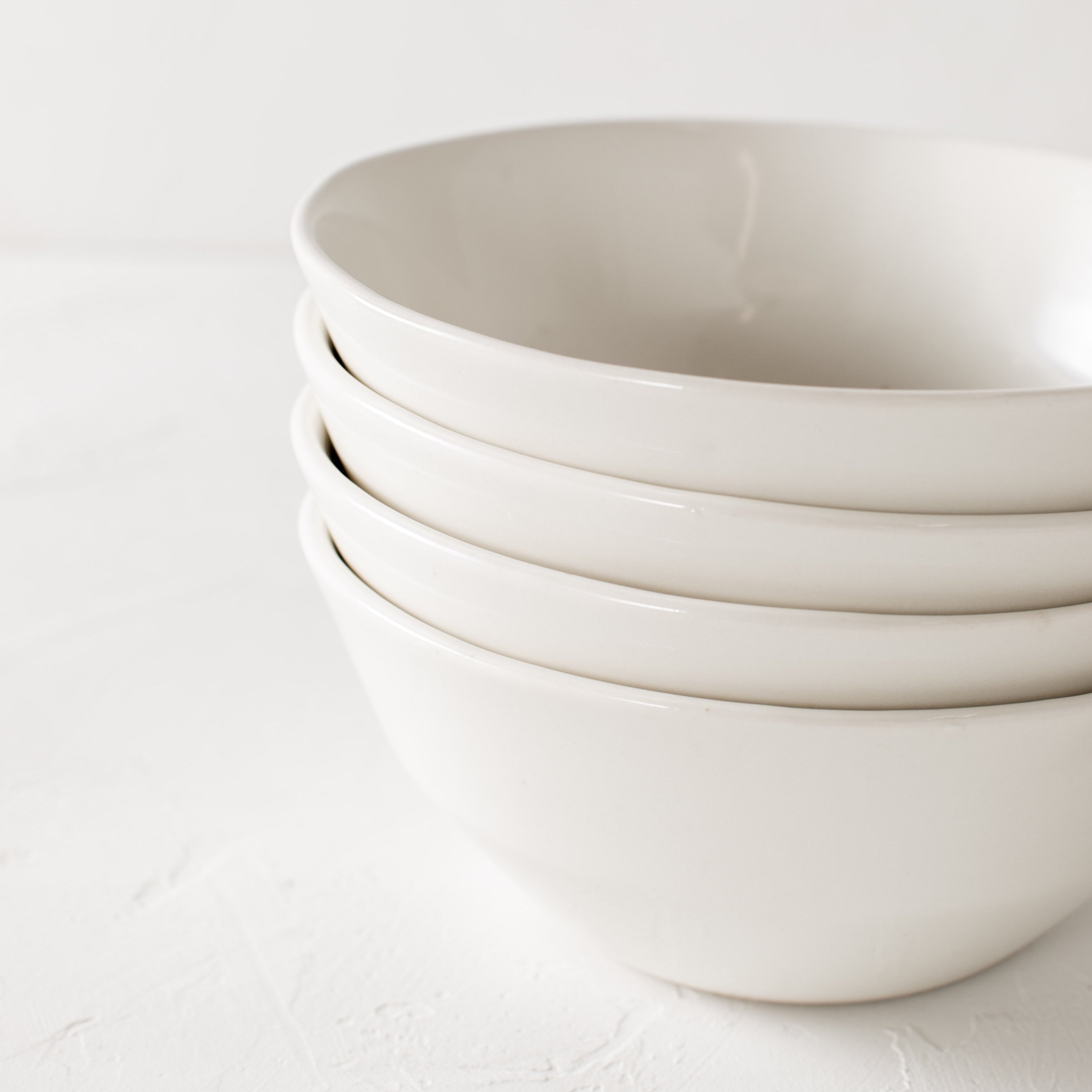 Four white ceramic minimal bowls staged in close up photo to show detail of bowl rim. Staged  on a white textured tabletop against a white textured backdrop. Handmade bowls are designed and sold by Convivial Production. Kansas City Ceramics.