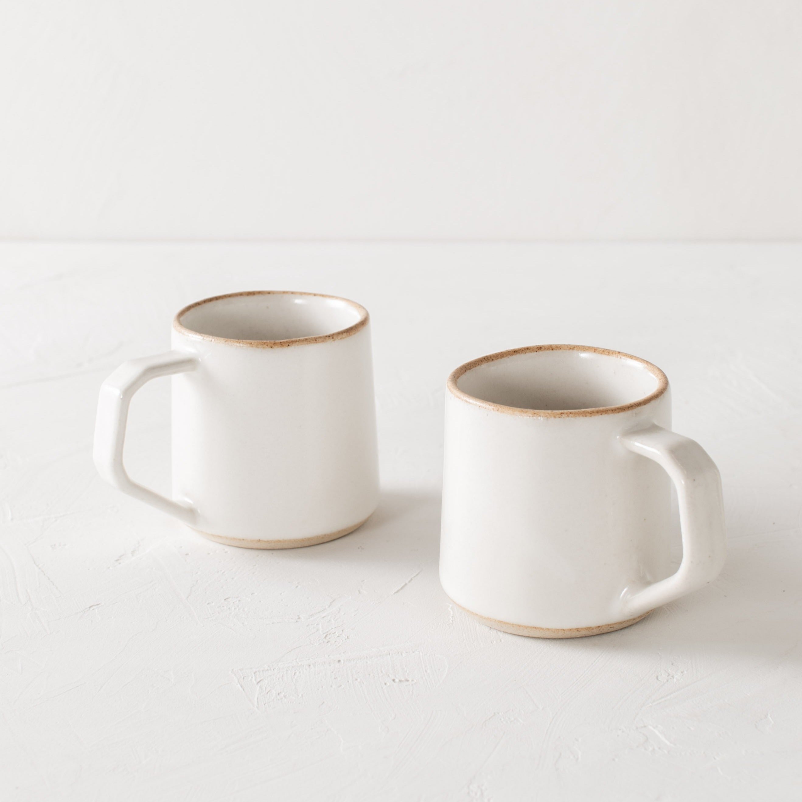 Pair of white minimal ceramic mug is staged on a white textured table top and white textured back drop. Mugs have exposed warm stoneware on the rims as well as the base. Handles are geometric in shape but still rounded. Convivial Production, Kansas City Ceramics. Handmade ceramic mug.