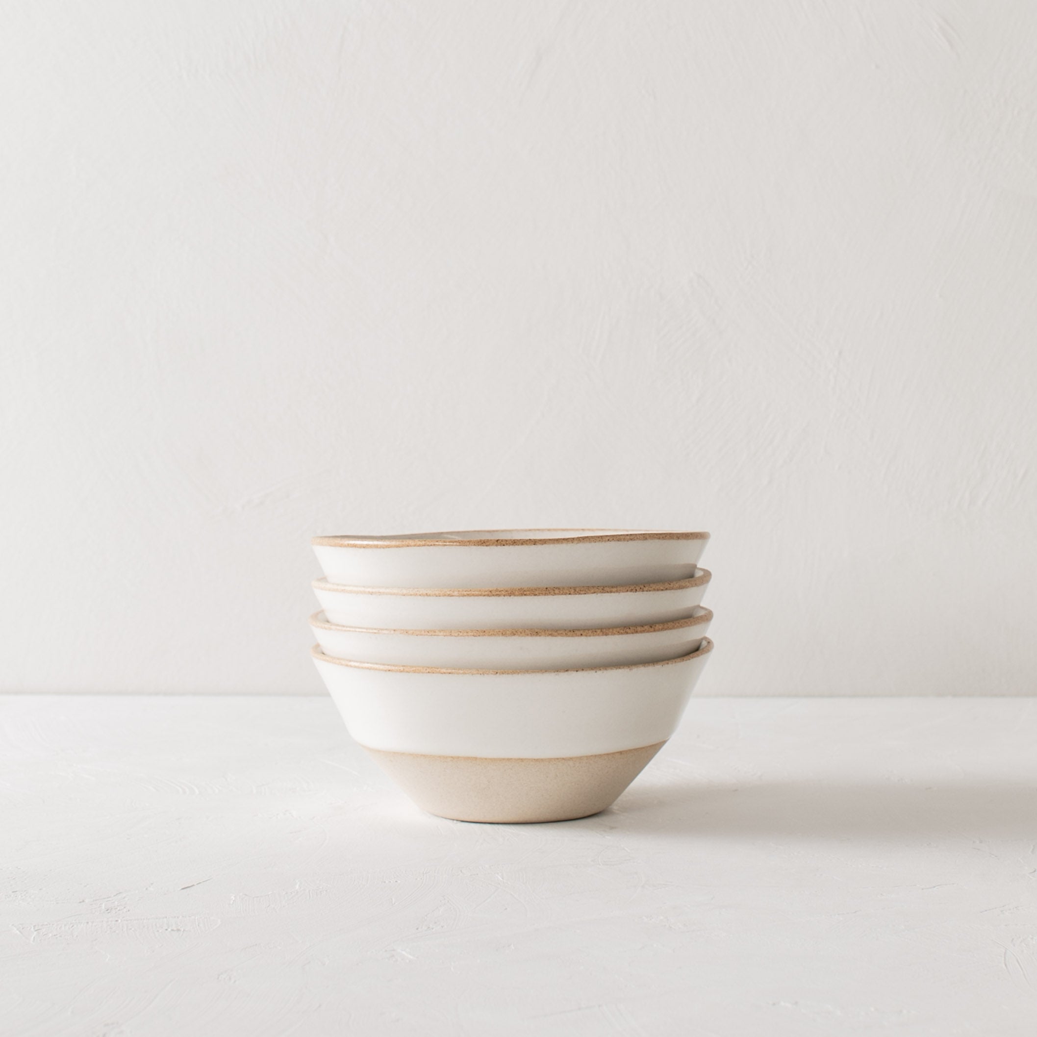 White ceramic bowls. Handmade glazed stoneware bowls, stacked together in a group of four. Bowls have exposed stoneware rims as well as bases. Centered in the image staged against a white textured wall and table top. Designed and sold by Convivial Production, Kansas City Ceramics.