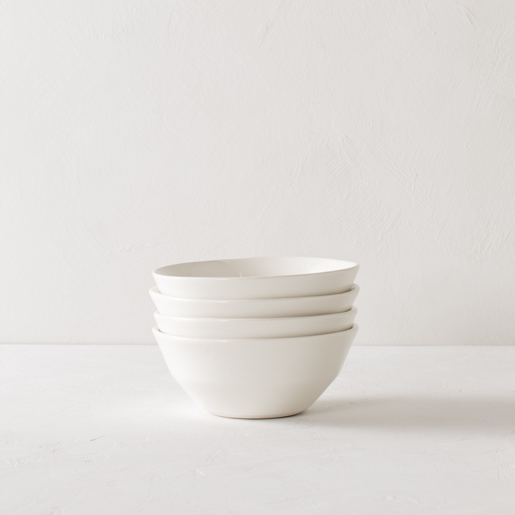 Four white minimal ceramic bowls stacked in the center on a white textured tabletop against a white textured backdrop. Handmade ceramics designed and sold by Convivial Production. Kansas City Ceramics.