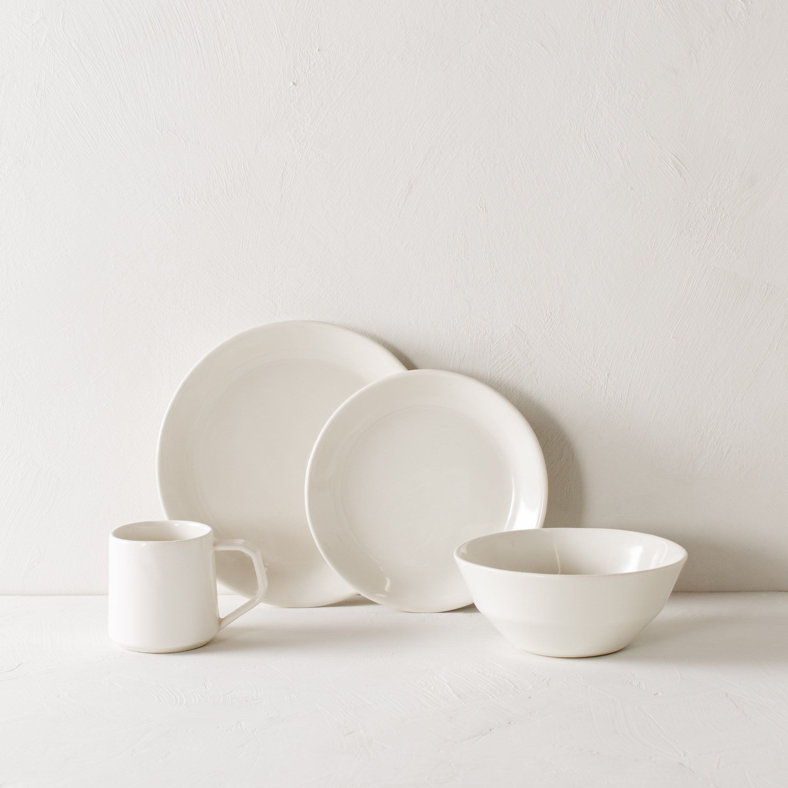 White minimal ceramic dinner set. Large and small plate leaning against white plaster wall. White ceramic mug and minimal bowl on each side. Handmade ceramics designed and sold by Convivial Production, Kansas City Ceramics.