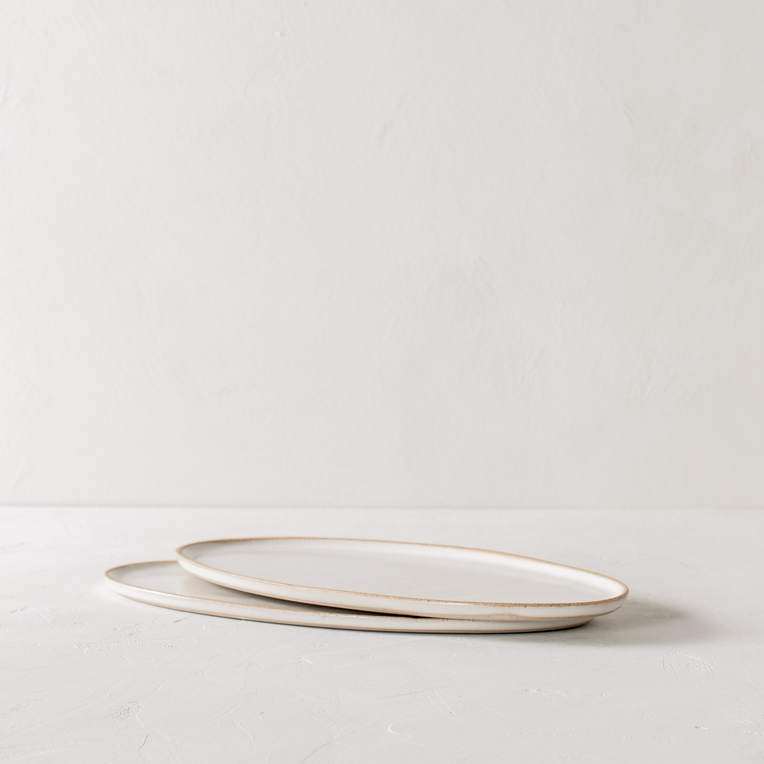 Two narrow vertical ceramic oval serving trays staggered and overlapping on a white textured table-top. Handmade ceramic serving tray, designed and sold by Convivial Production, Kansas City ceramics.